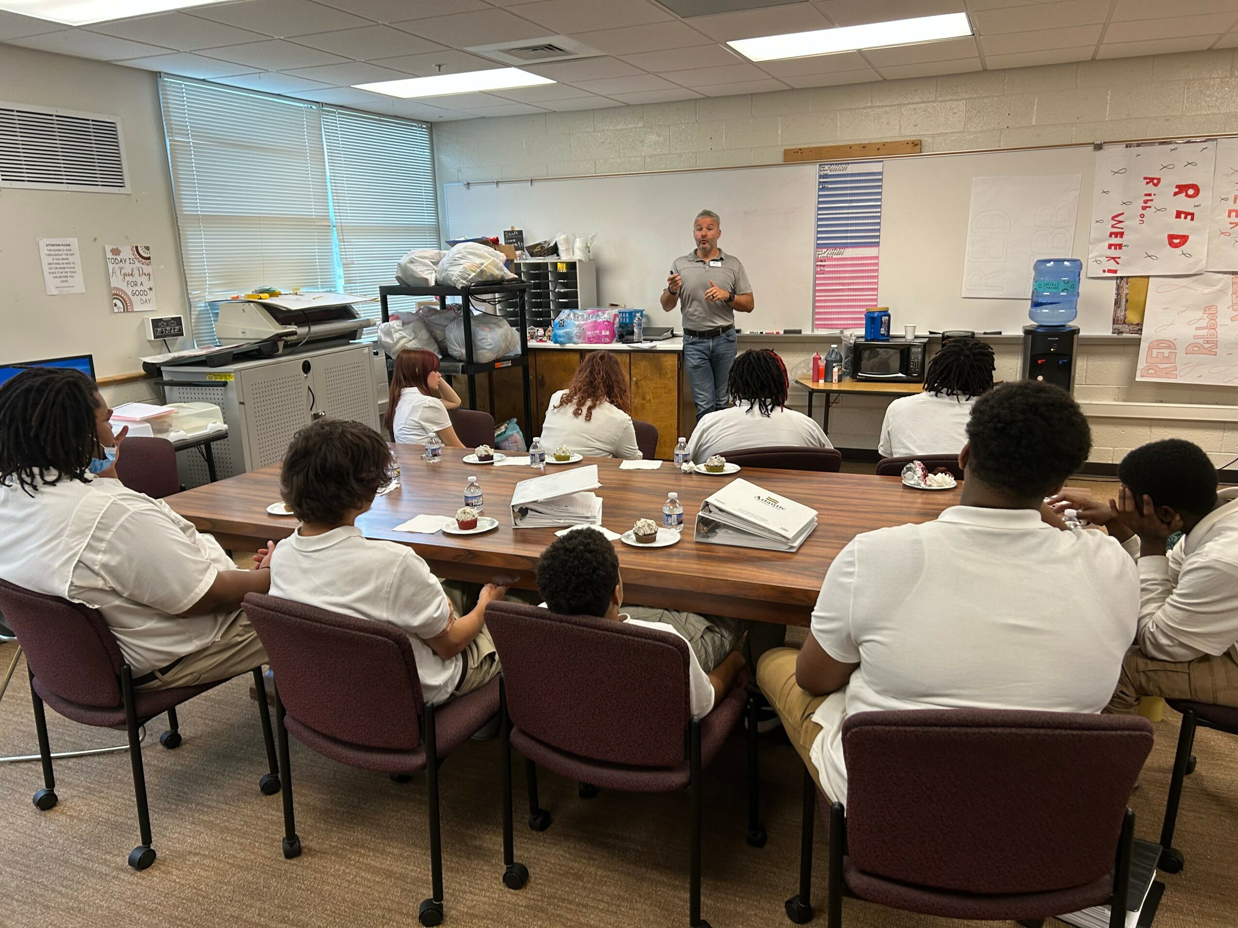 Teacher standing at front of classroom with students seated at tables, some wearing white uniforms, in a classroom setting.