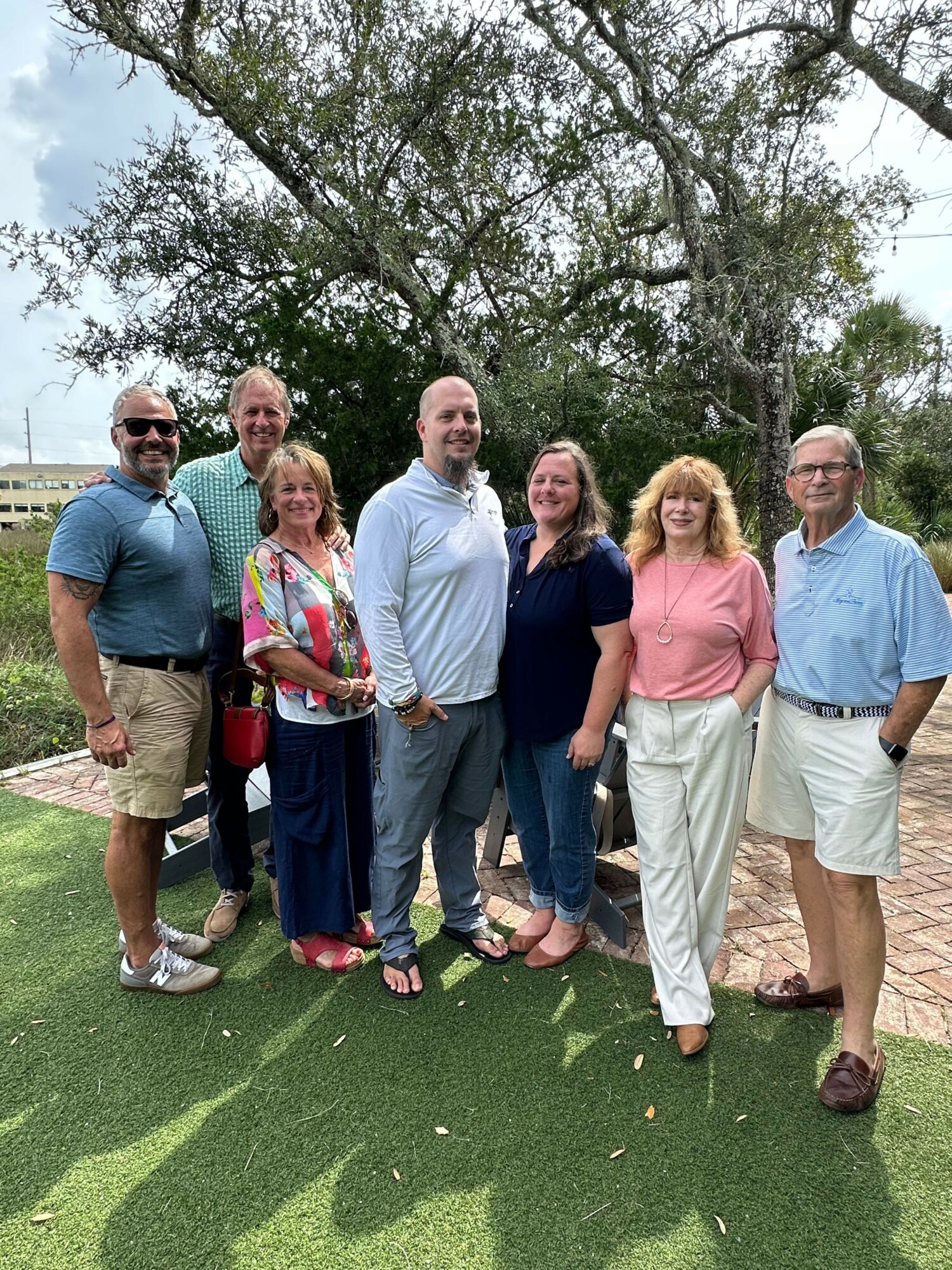 Group of seven people standing outdoors on grass, smiling, with trees and sky in background.
