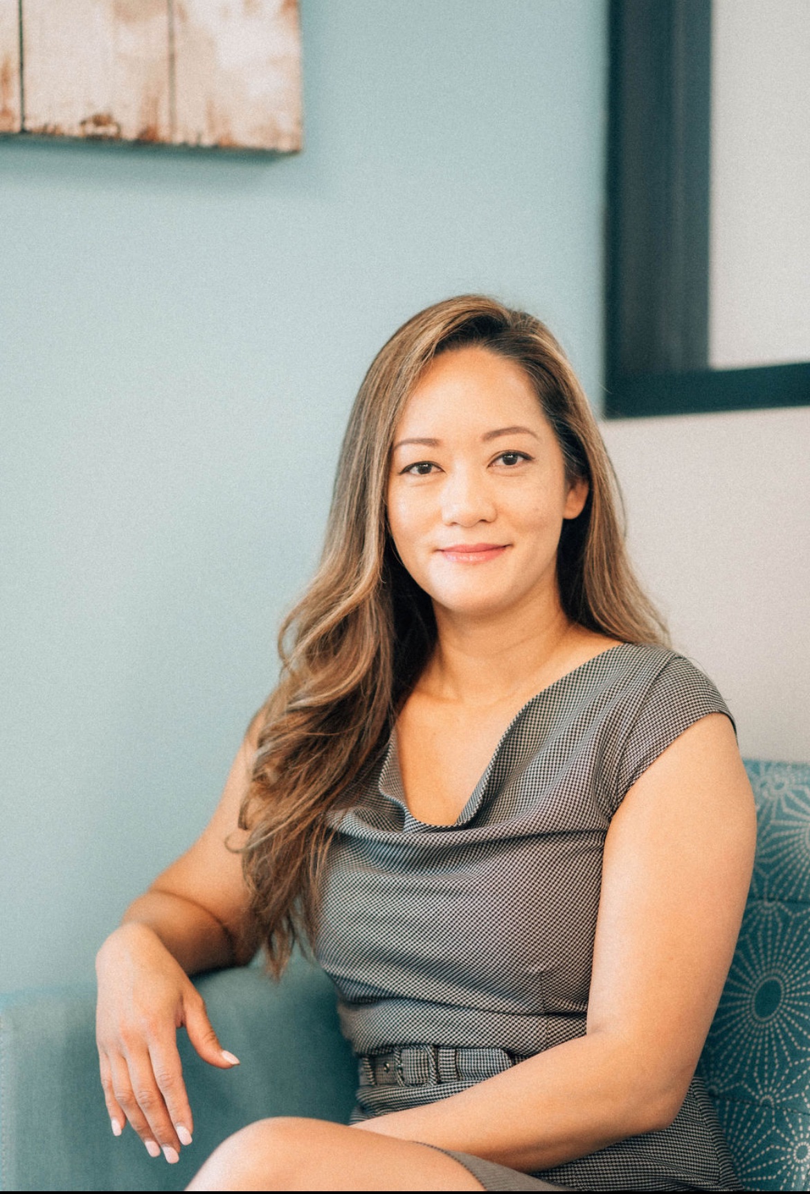 Woman with long hair sitting on a chair, smiling, in a room with light blue wall and window.
