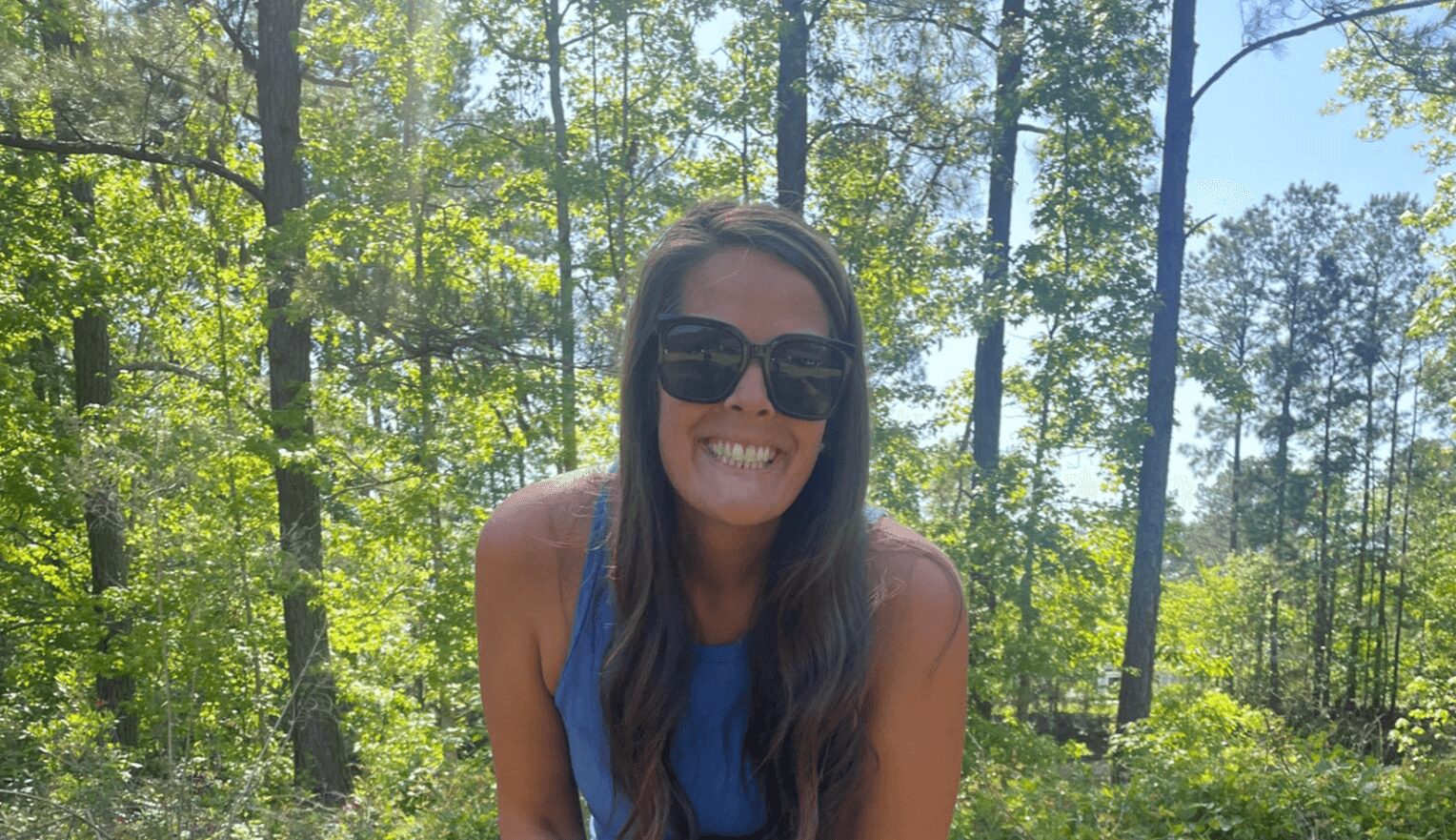 Smiling woman with long hair and sunglasses outdoors in a wooded area.