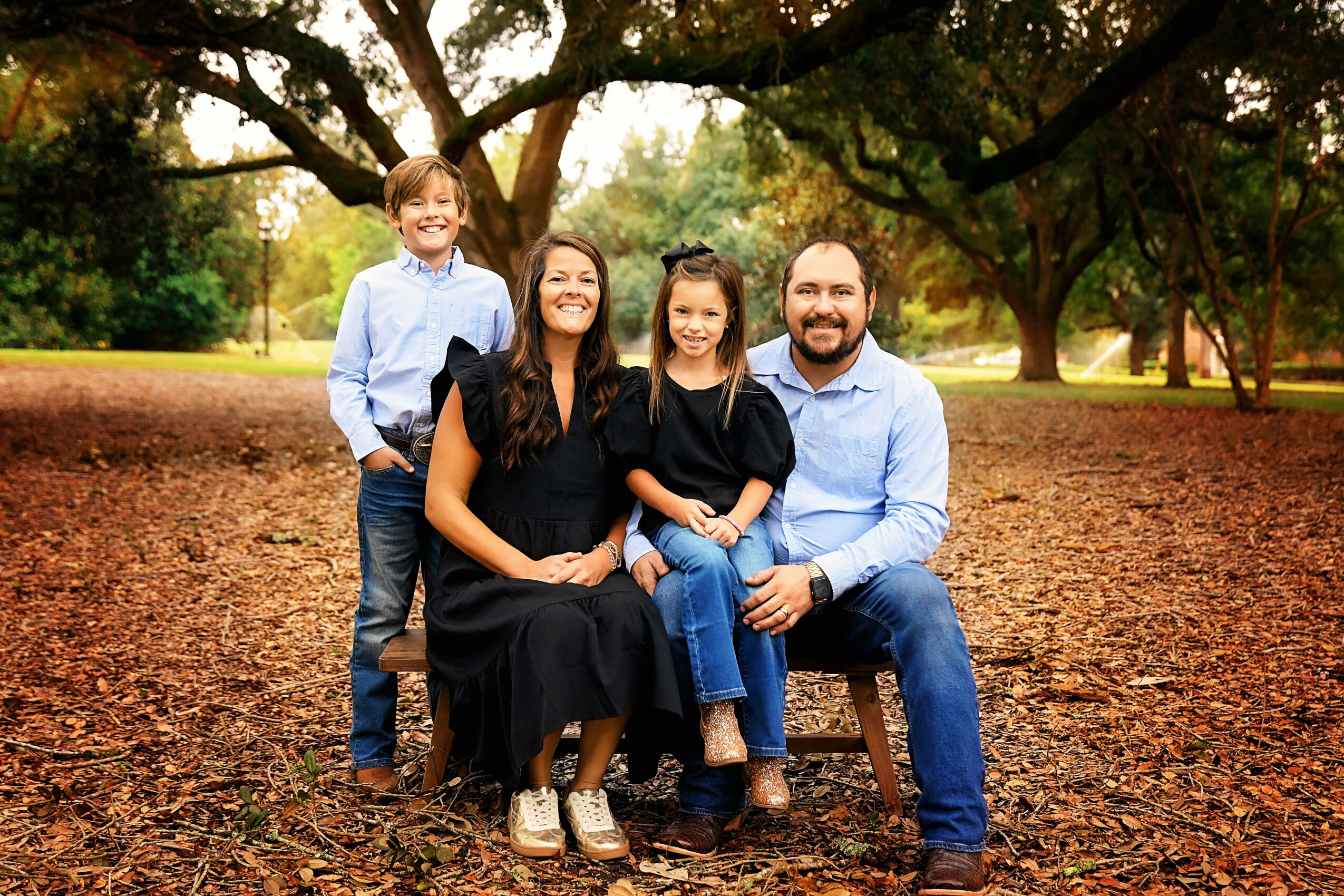 Family of four sitting and standing outdoors on a dirt ground with trees in background, smiling at camera.