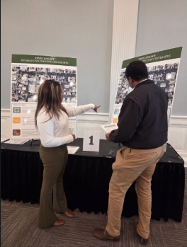 Two people standing at a table with display boards, one pointing at the board, the other holding a paper, in an indoor setting.