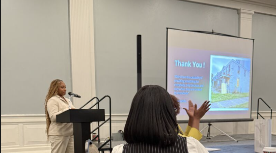 Woman speaking at podium in front of a presentation screen, audience member raising hand, plain wall background.