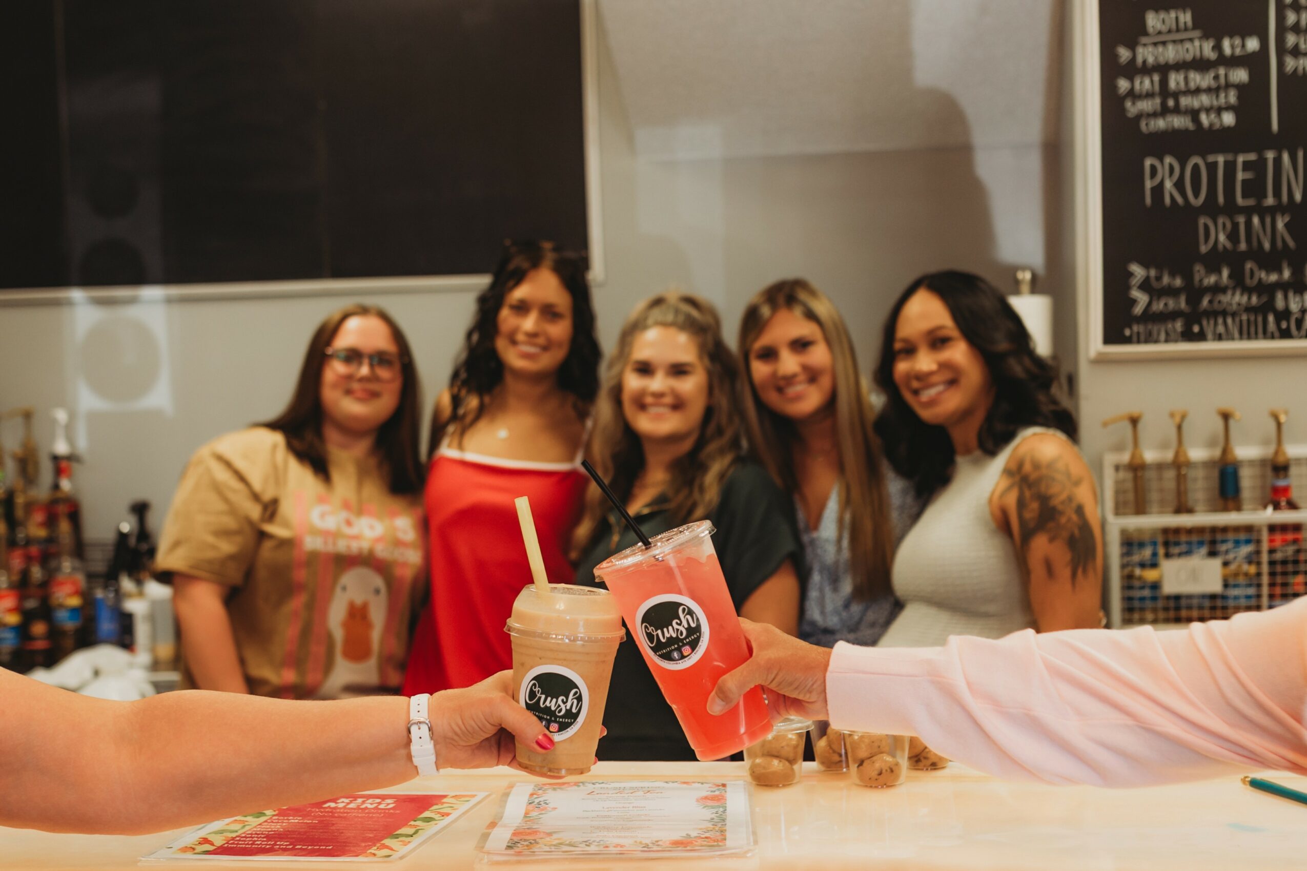Five women smiling and holding drinks in a bar or cafe setting, with a chalkboard menu in the background.
