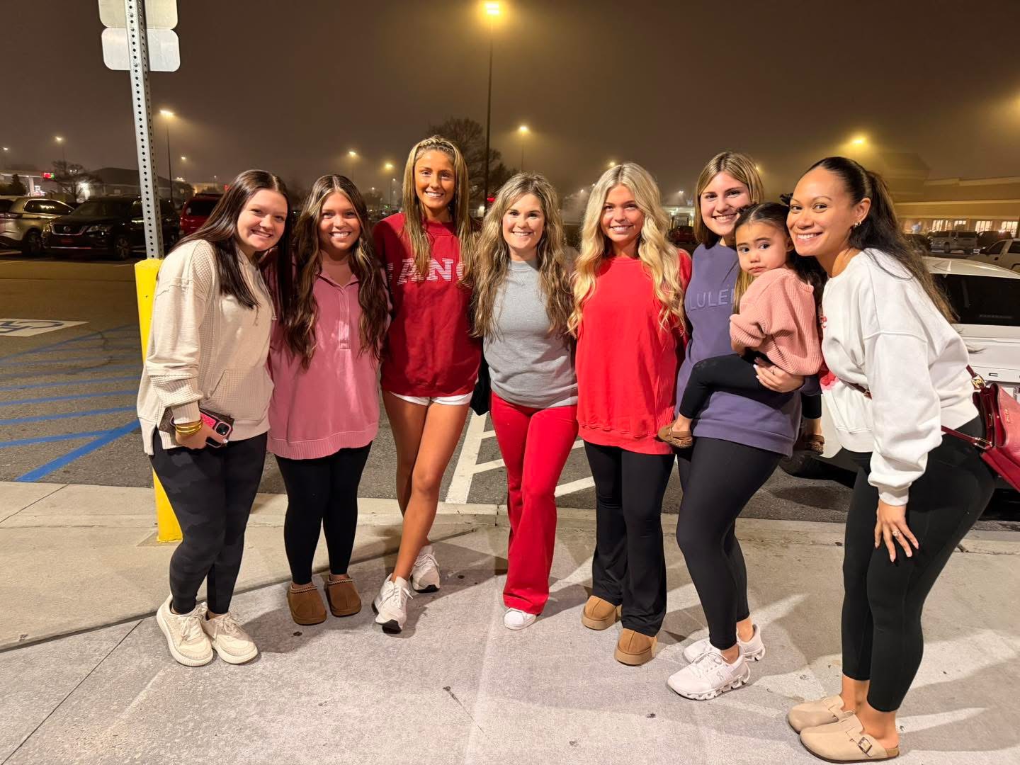 Group of eight women and children standing outdoors at night, smiling, with cars and streetlights in background.