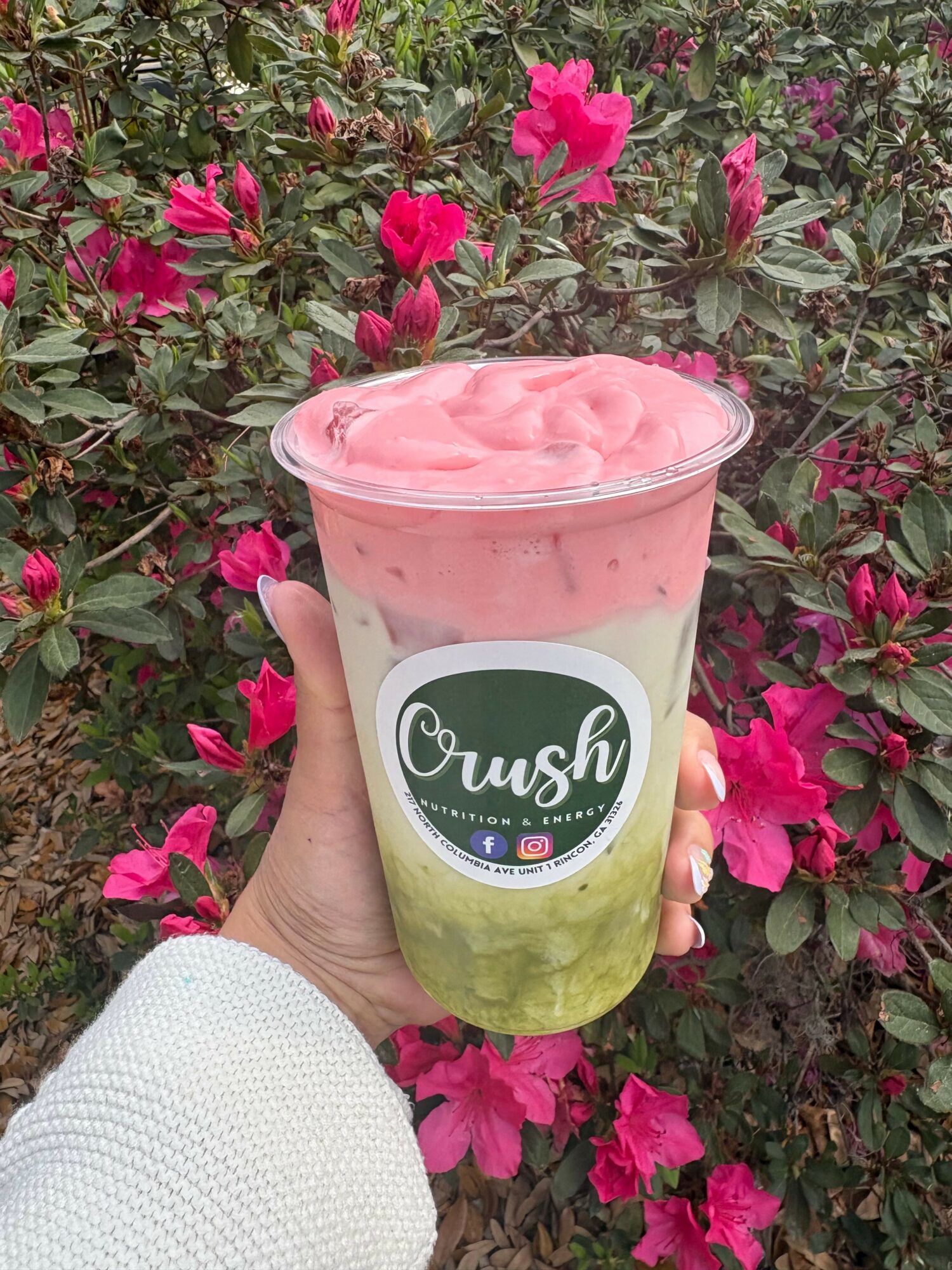Hand holding a layered drink with pink foam, green jelly, and white base in front of pink flowers.
