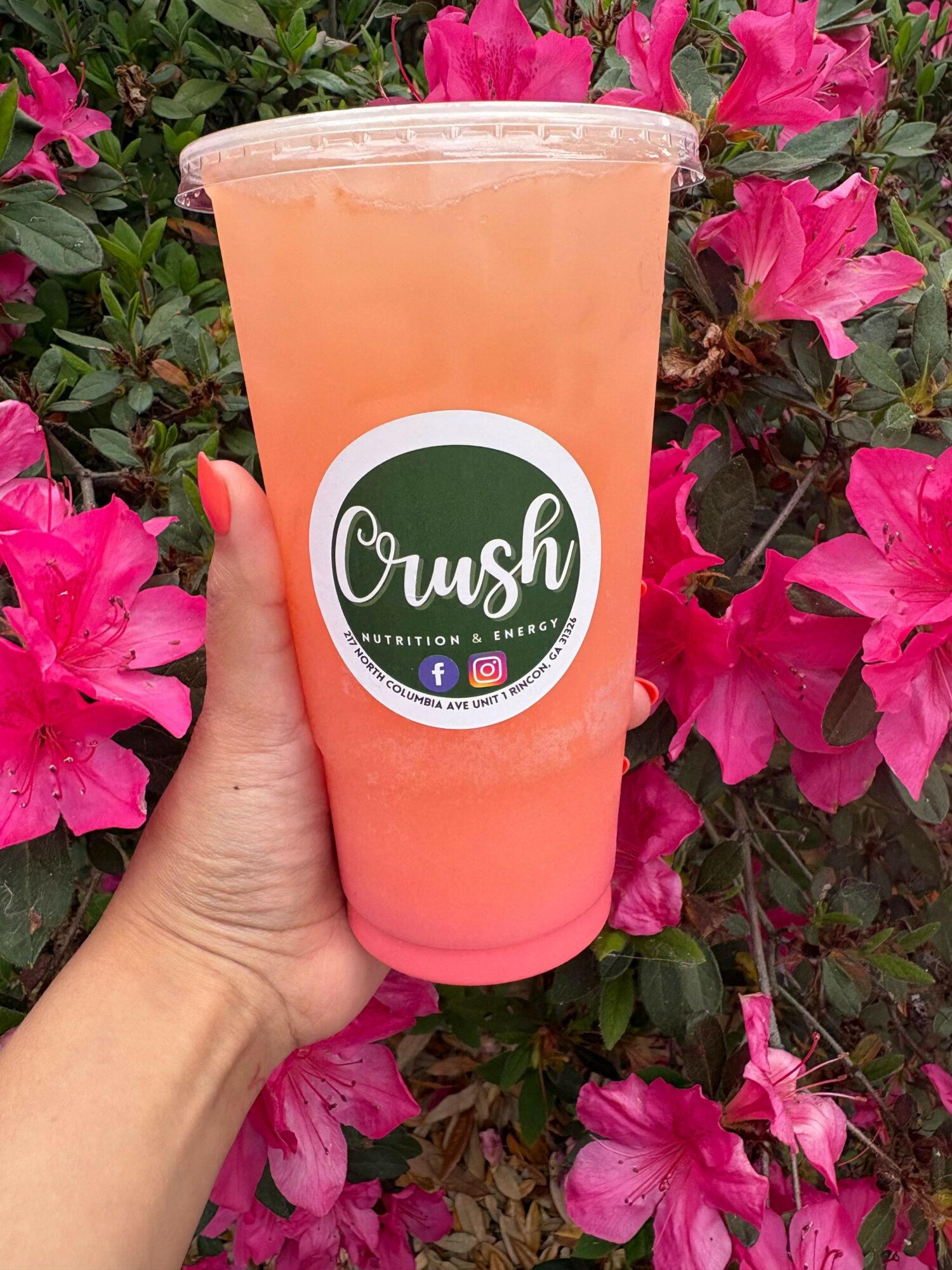 Person holding a pink-colored drink in a clear cup with a Crush logo, surrounded by pink flowers.