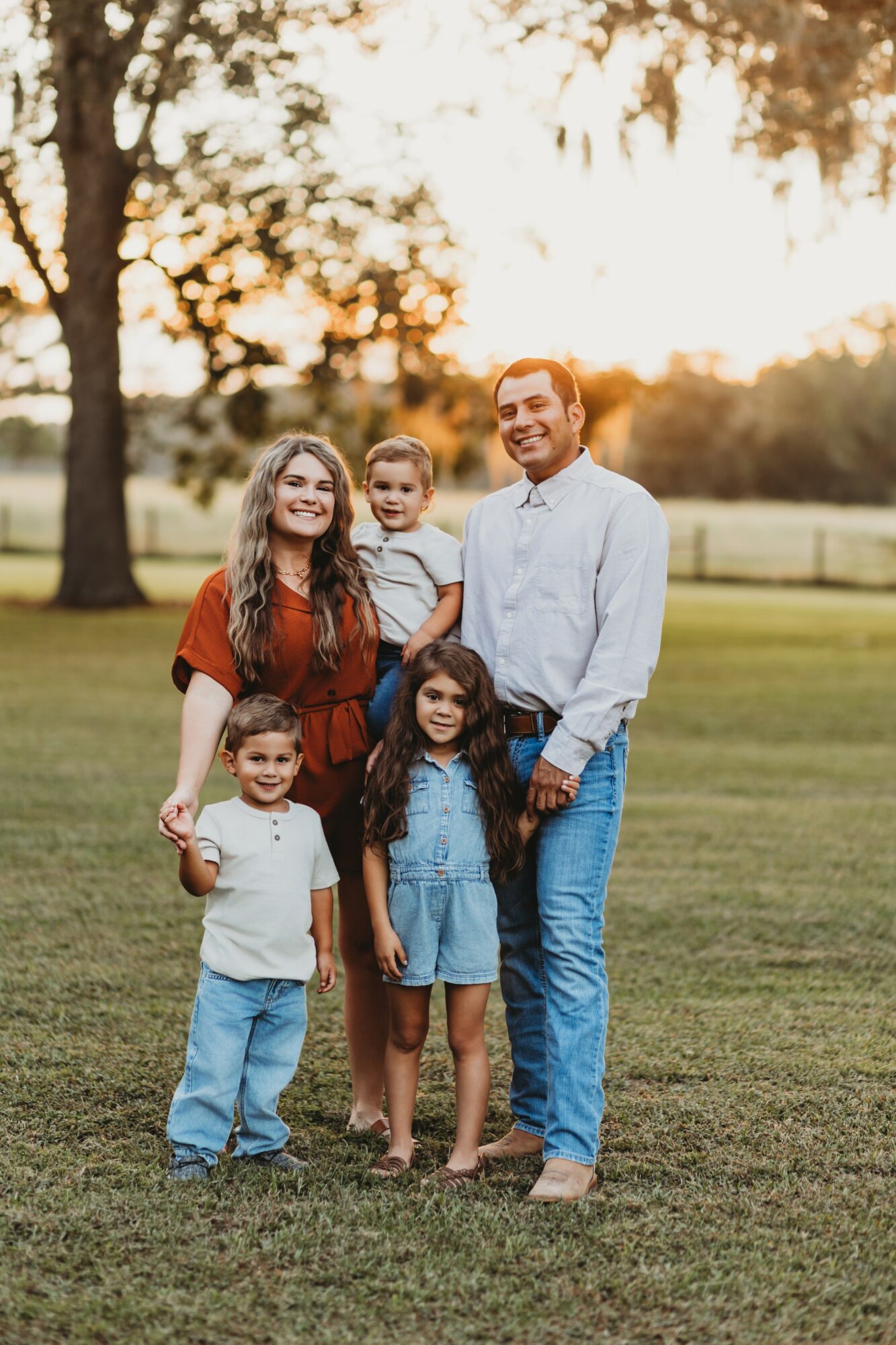 Family of five standing outdoors on grass during sunset, smiling, with trees and a fence in background.