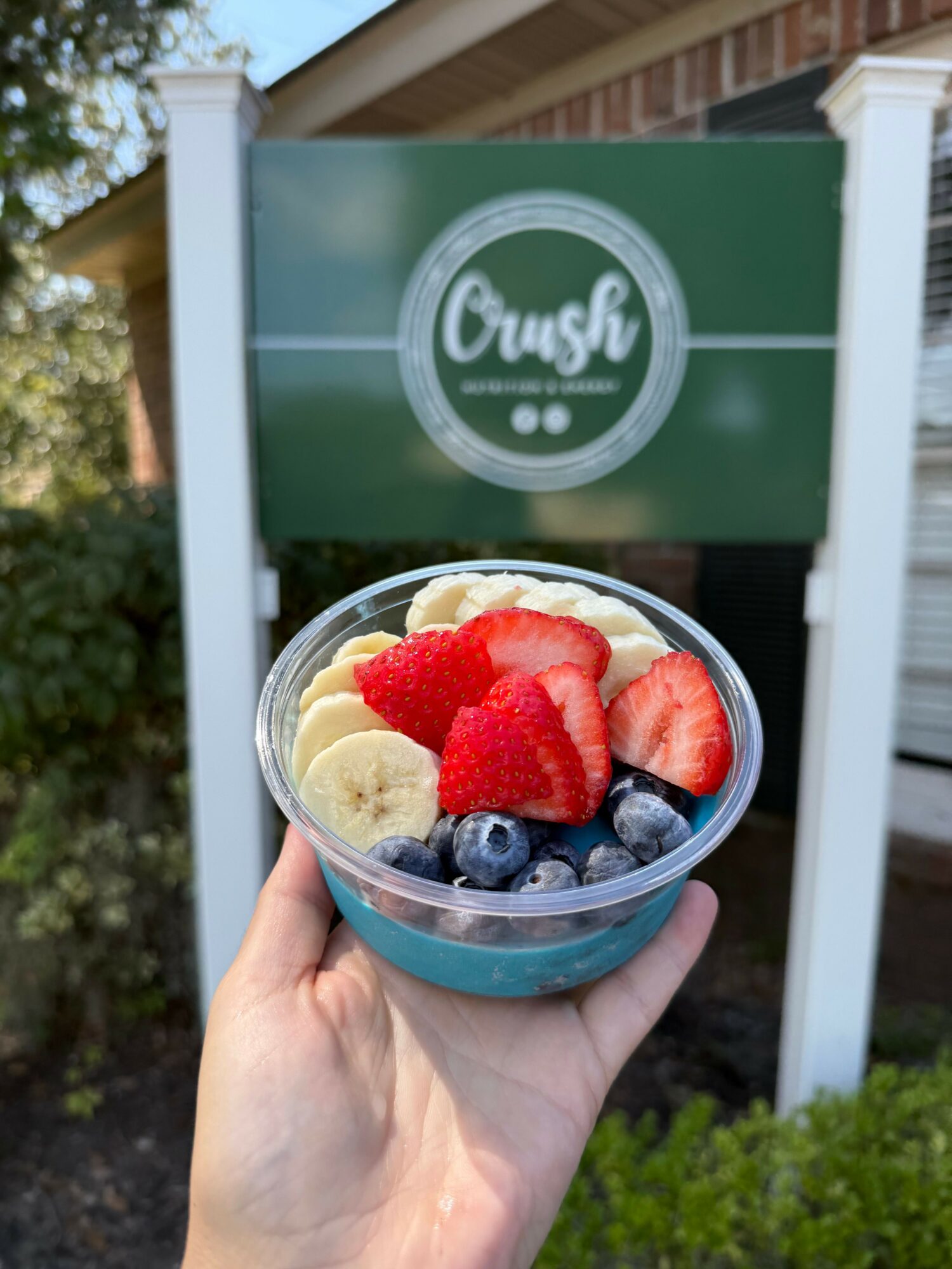 Hand holding a cup of smoothie with bananas, strawberries, and blueberries in front of a green Crush sign.