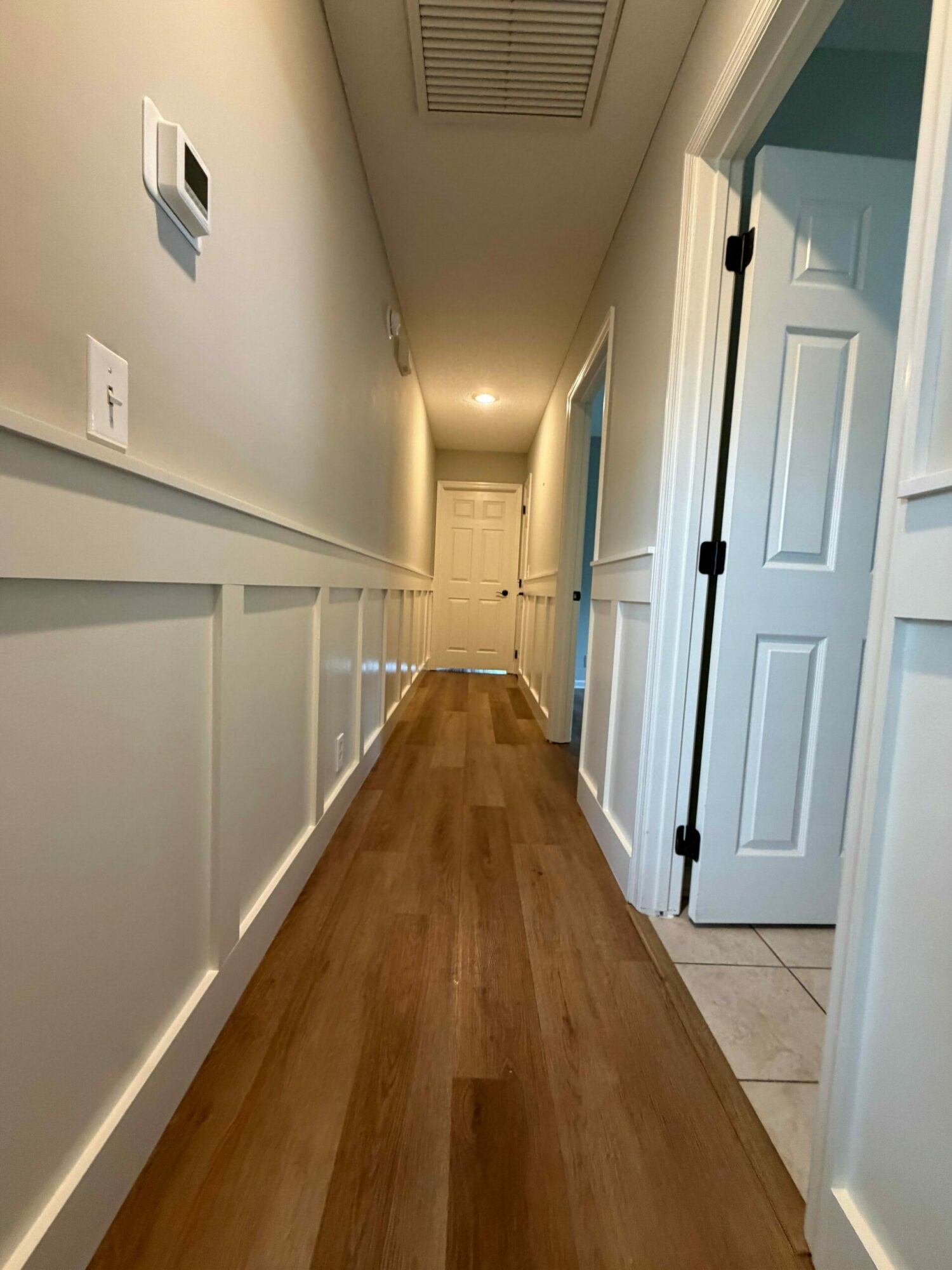 Long hallway with white walls, a wooden floor, and a door at the end, illuminated by ceiling lights.