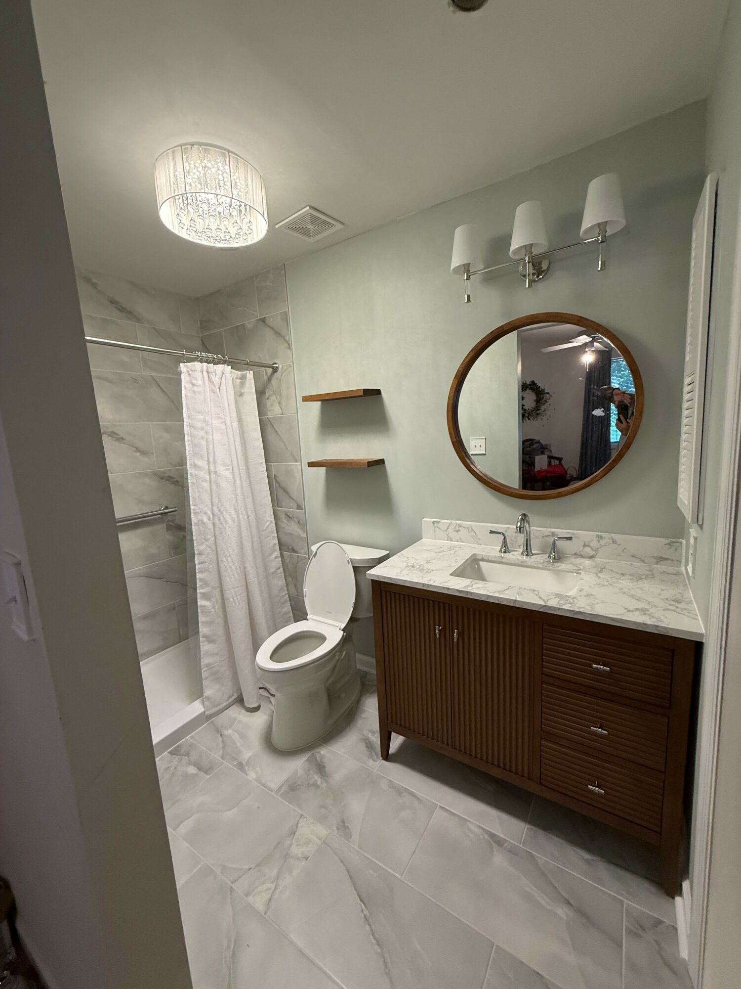 Bathroom with shower, toilet, wooden vanity with marble countertop, round mirror, wall-mounted lights, and shelves, light-colored walls and floor.