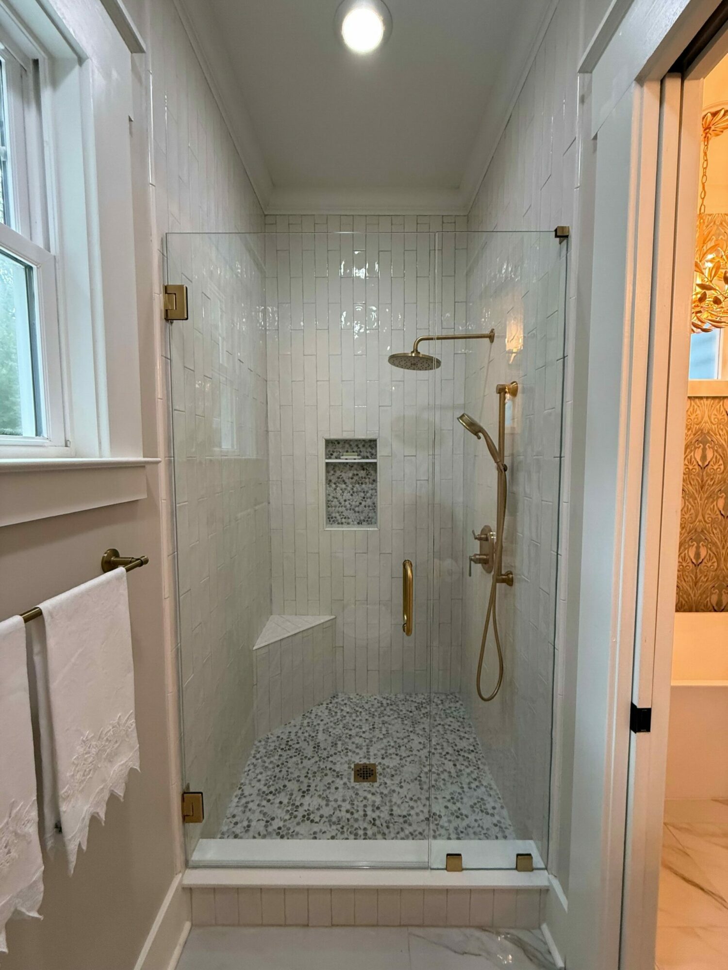 Bathroom shower with white tiled walls, a showerhead, and a small window, viewed through a glass door.