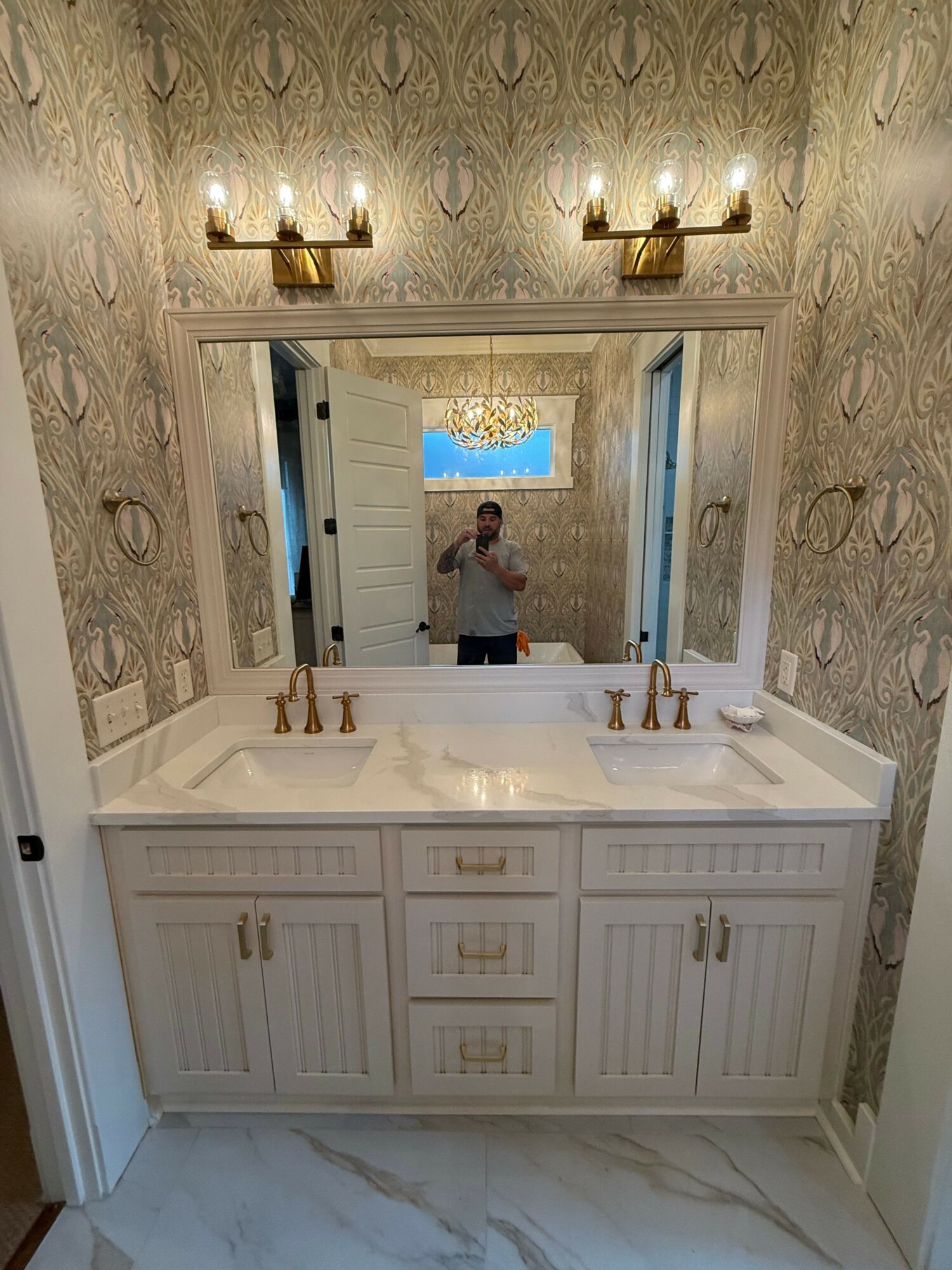 Bathroom vanity with two sinks, large mirror, and decorative wallpaper, person taking photo reflected in mirror.