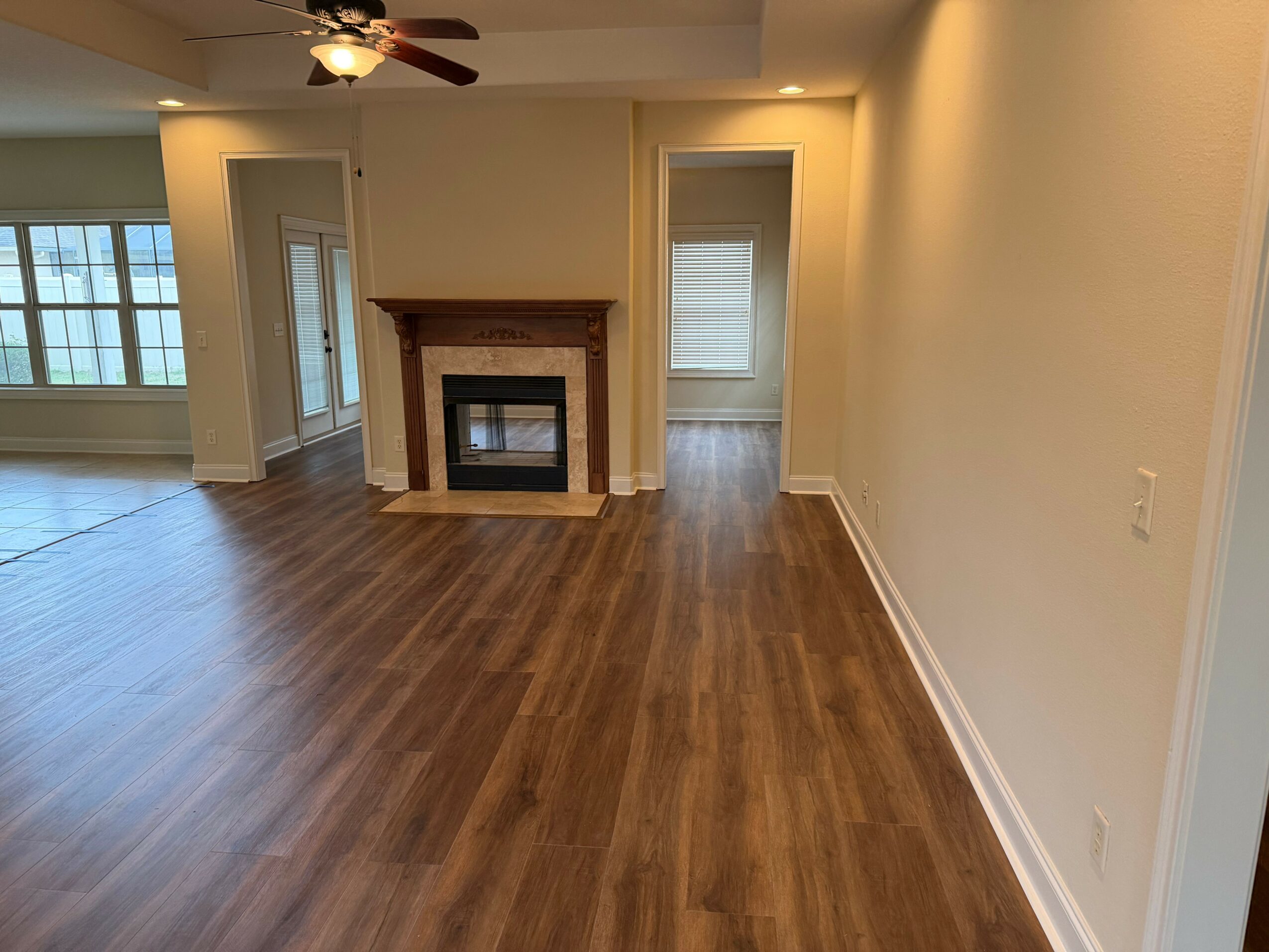 Empty living room with hardwood floor, fireplace, ceiling fan, and windows, with doorways leading to other rooms.