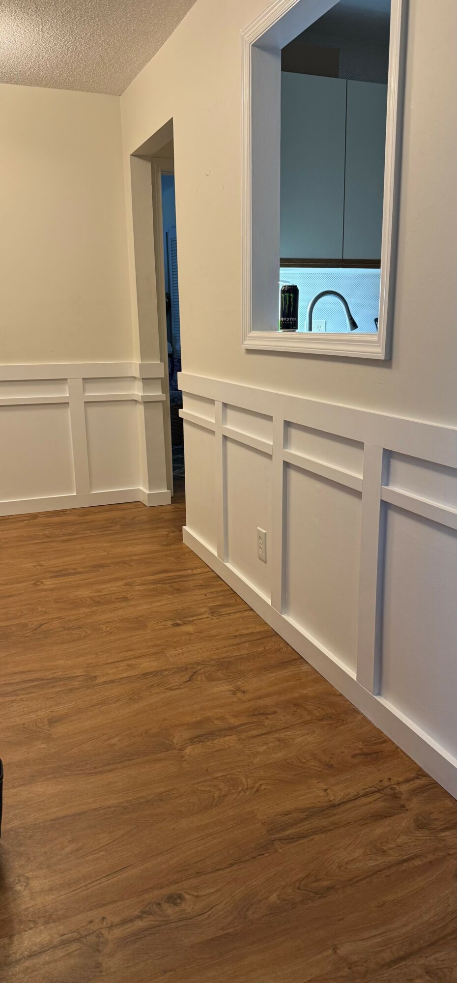 Interior wall with white wainscoting and a window looking into a kitchen with a faucet.