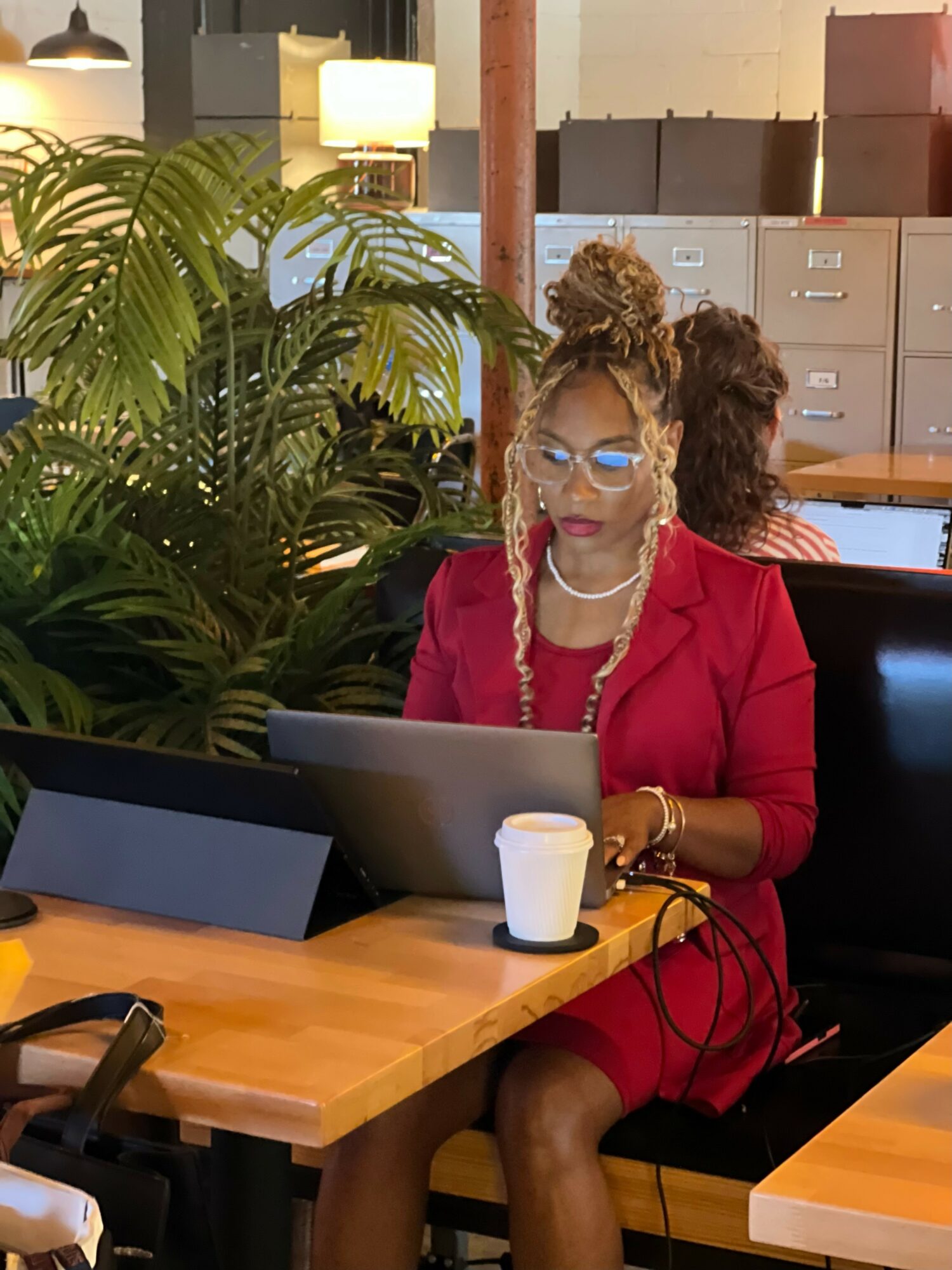 Woman with glasses and pearl necklace working on laptop at desk, large plant nearby, office background.
