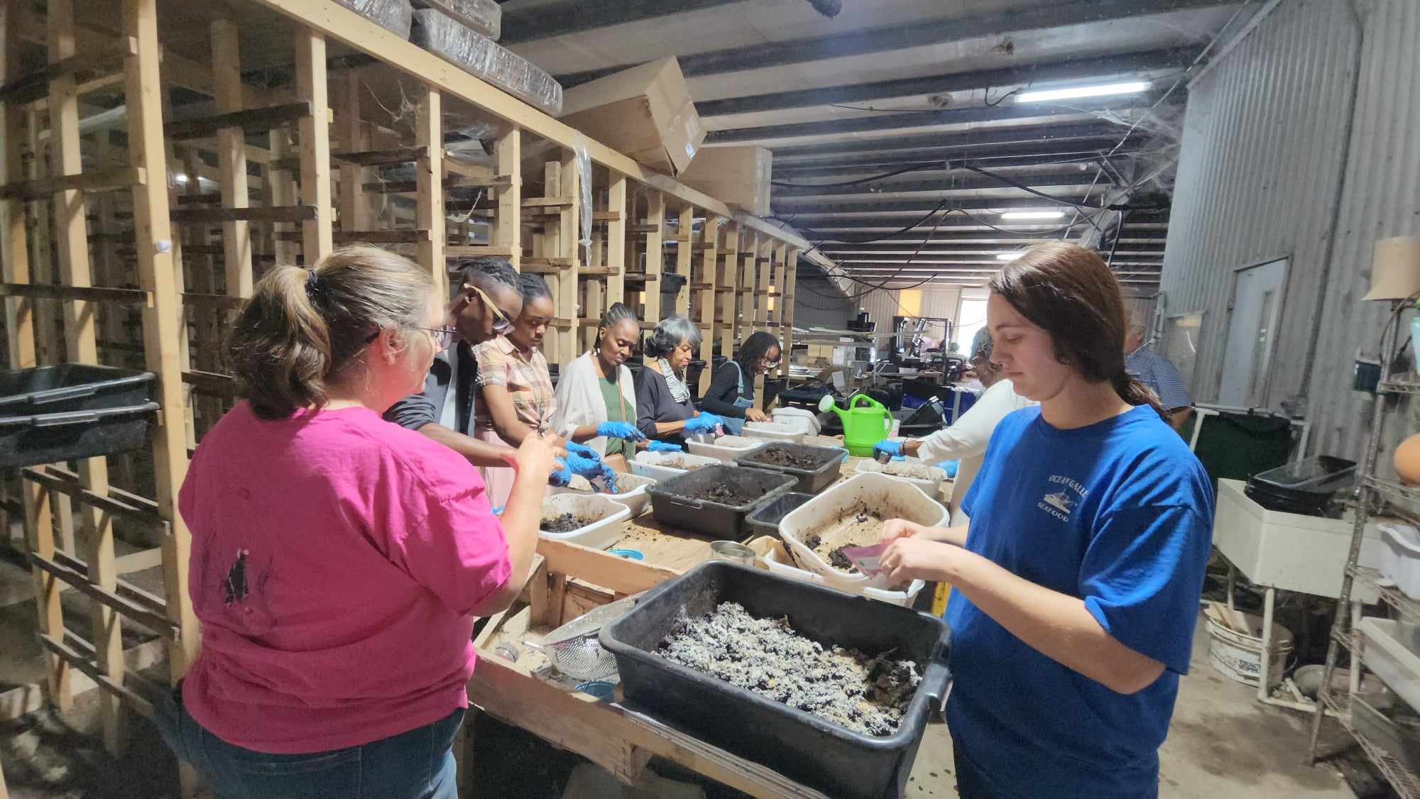 Group of people working in a warehouse or industrial space, handling materials at a long workbench.