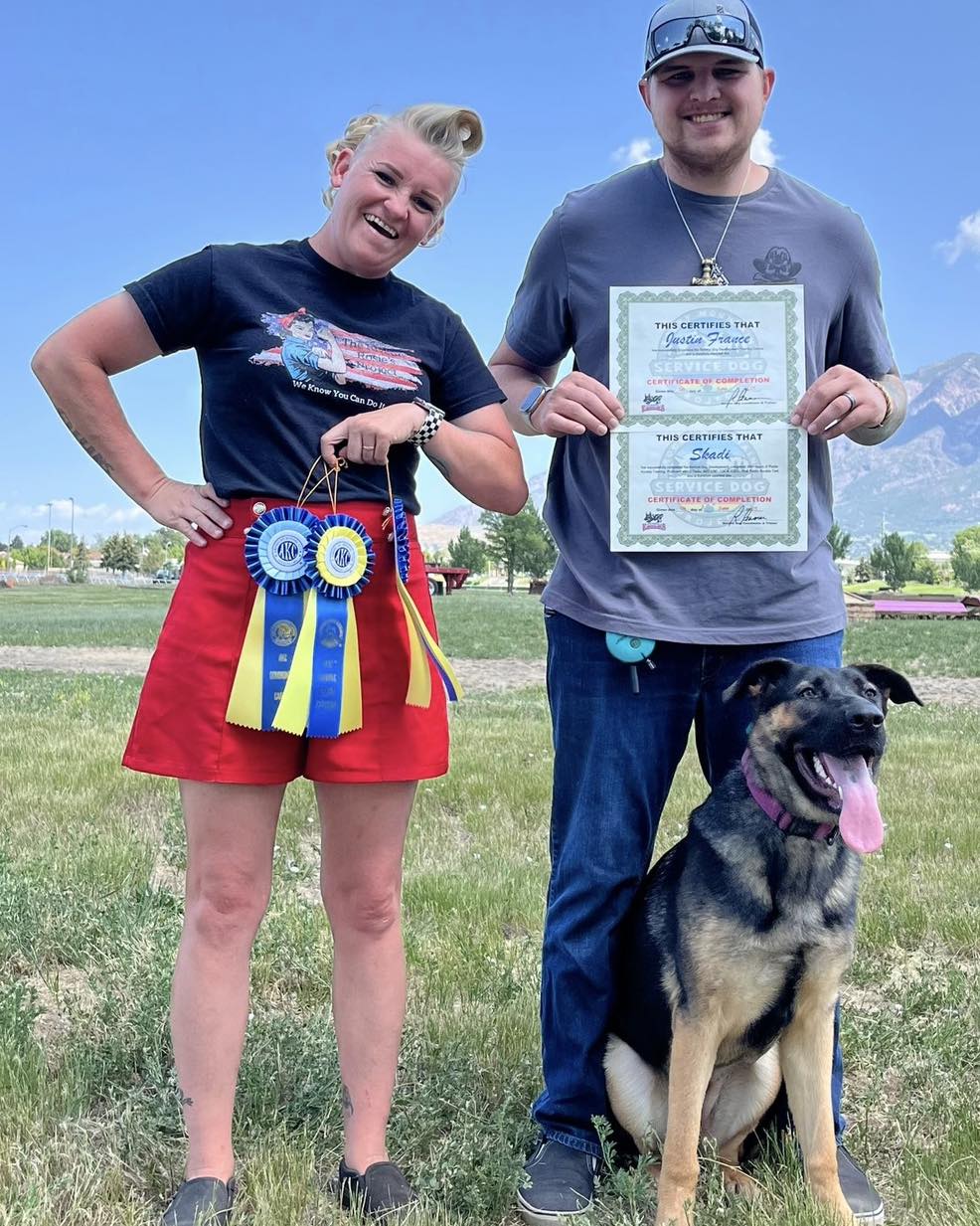 Two people and a dog outdoors, smiling, holding certificates, with mountains and blue sky in background.