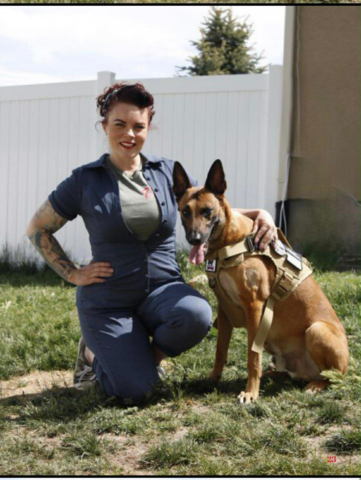 Woman kneeling outdoors with a Belgian Malinois dog wearing a harness, in front of a white fence and trees.