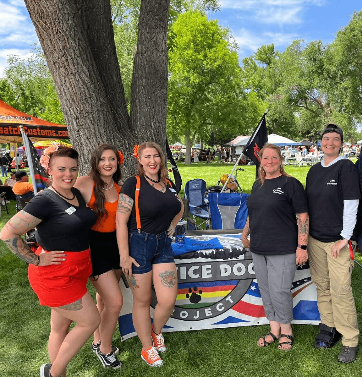 Group of five people standing outdoors near a table with a rainbow-colored sign, trees, and tents in background.