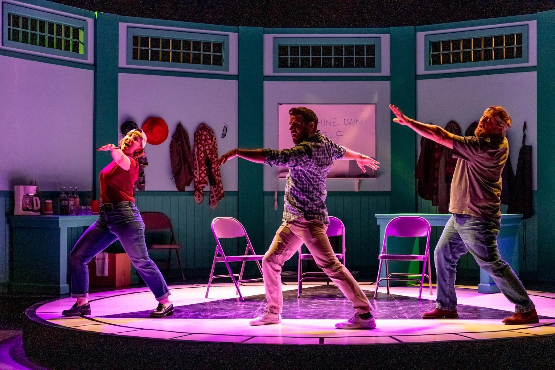 Three people dance on a stage with purple lighting, in a room with teal walls and chairs.