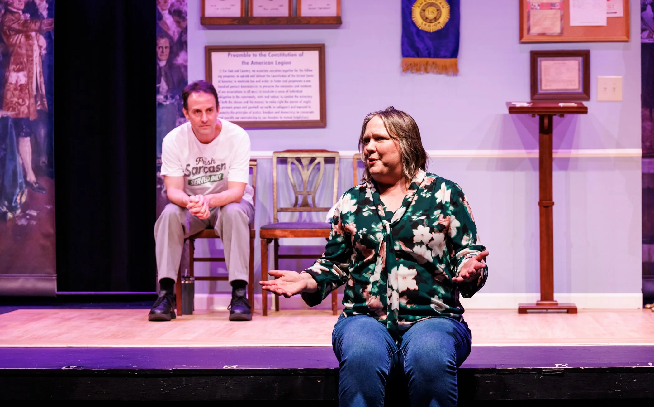 Two women on stage, one sitting and one standing, with purple walls and framed items in the background.
