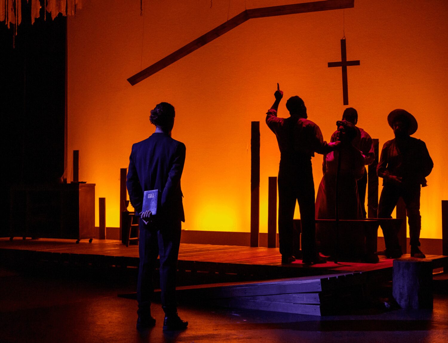 People on stage with a cross in the background, one person raising a finger, others standing nearby, dim lighting.