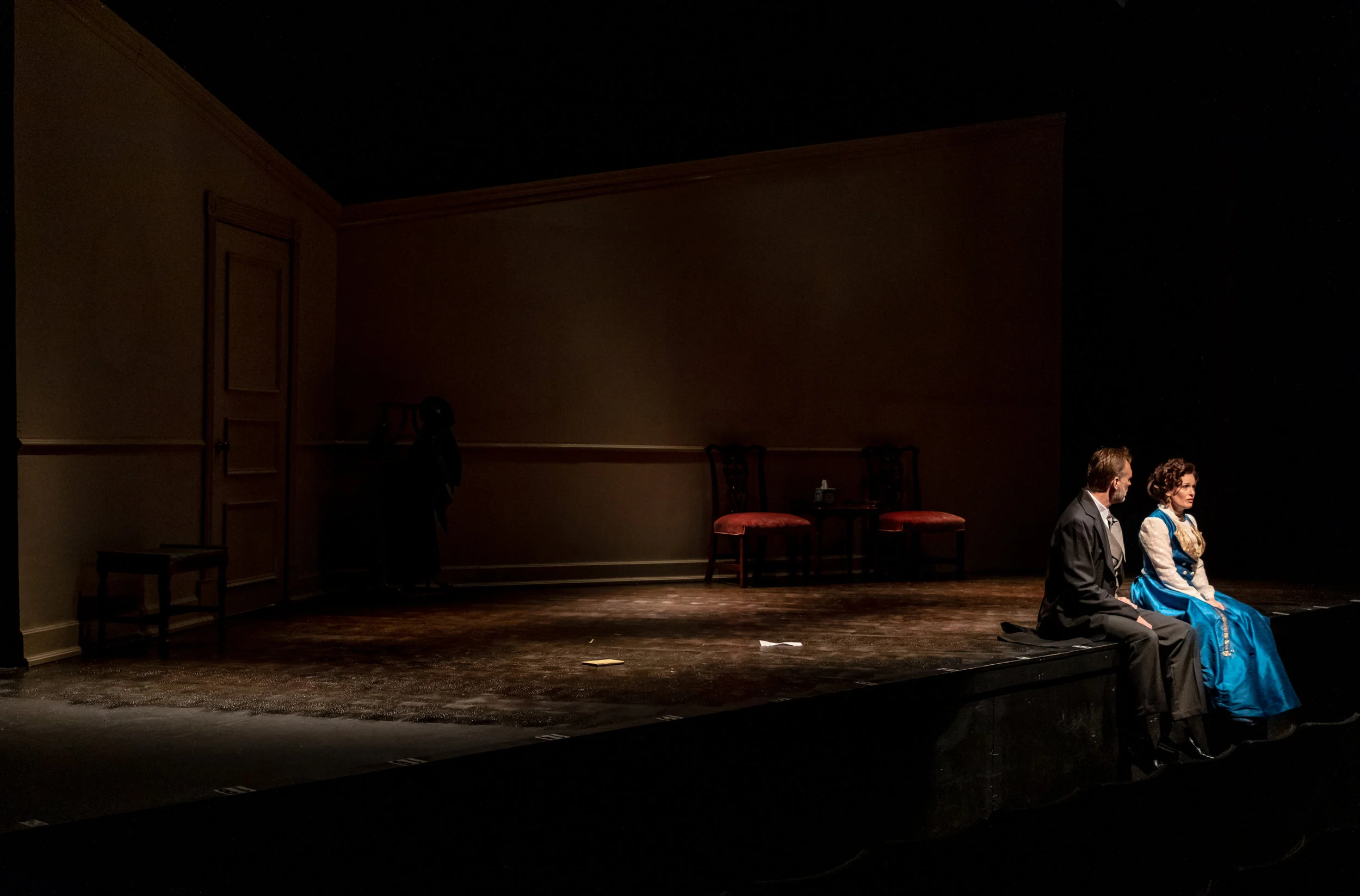 Two people sit on stage edge, with empty stage and furniture in background, dim lighting, and a closed door on the left.
