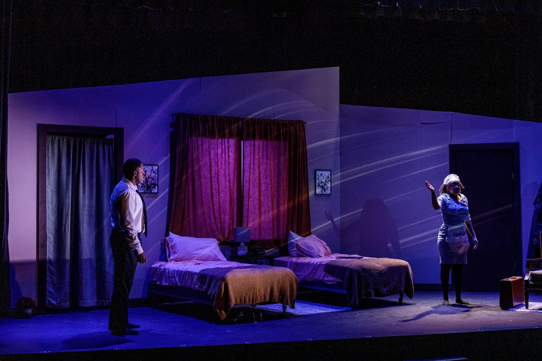 Stage scene with two beds, curtains, and three people, one speaking and two listening, in a theatrical setting.