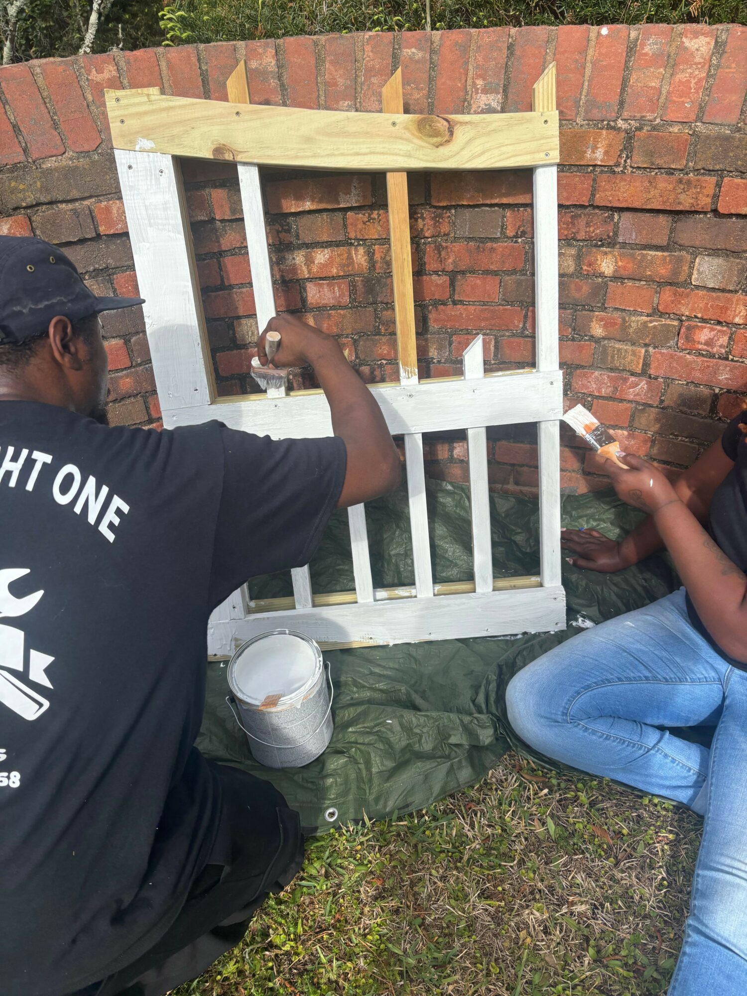 Two people painting a wooden fence panel outdoors, with a brick wall in the background. One person is sitting on the ground, the other is kneeling.
