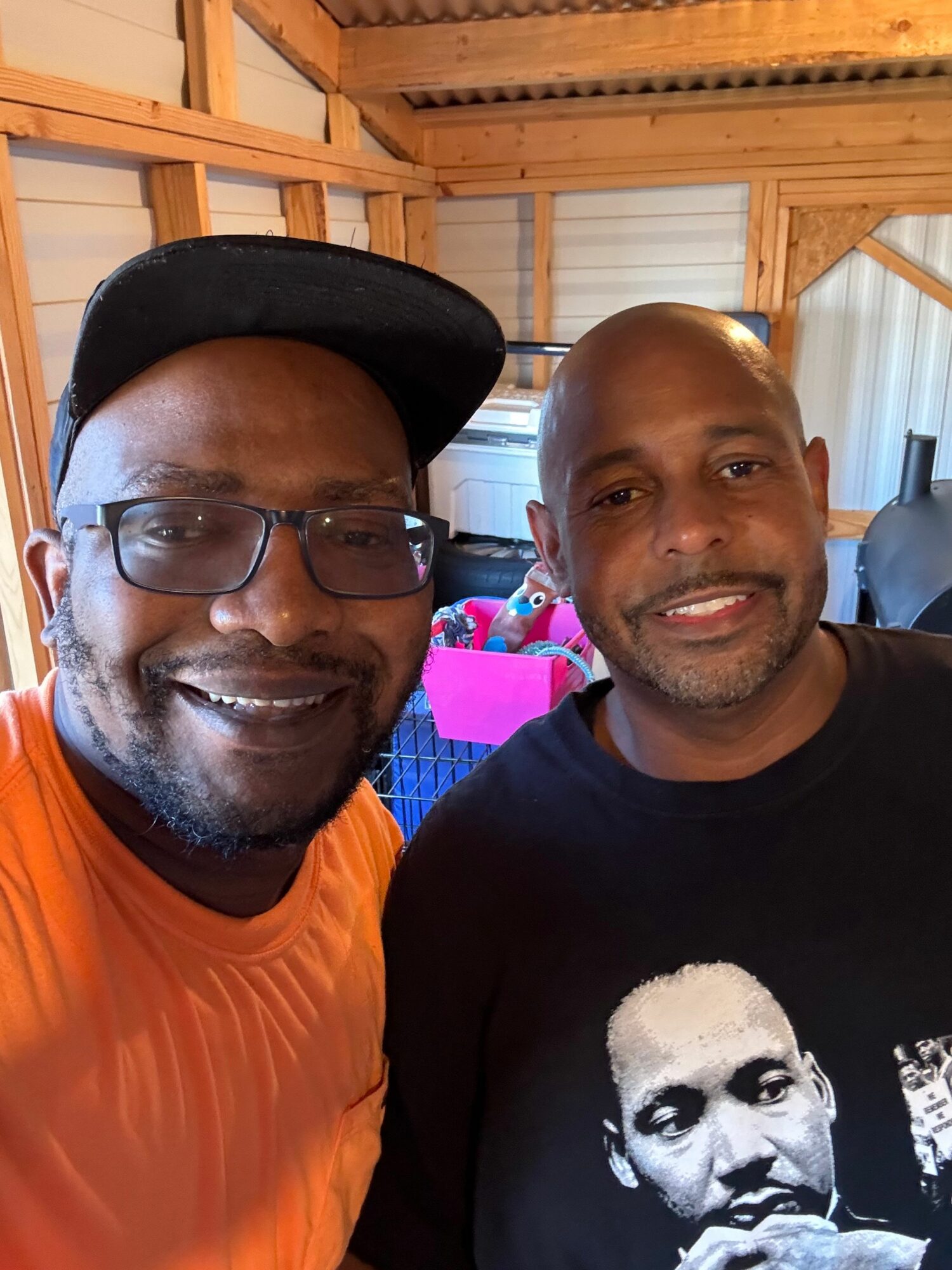 Two men smiling indoors, one wearing glasses and a cap, the other with a shaved head, in a wooden structure.