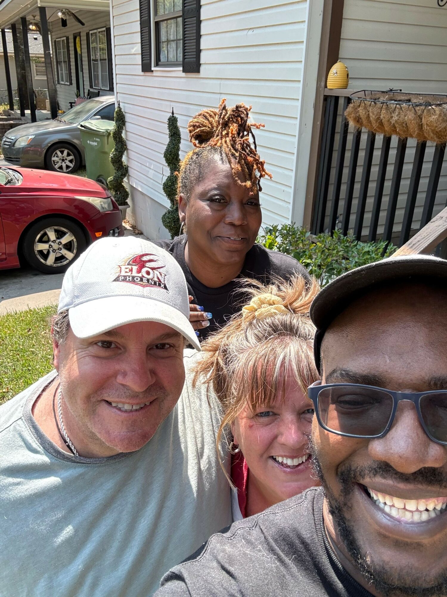Four people smiling outdoors in front of a house with a white exterior, cars, and greenery.