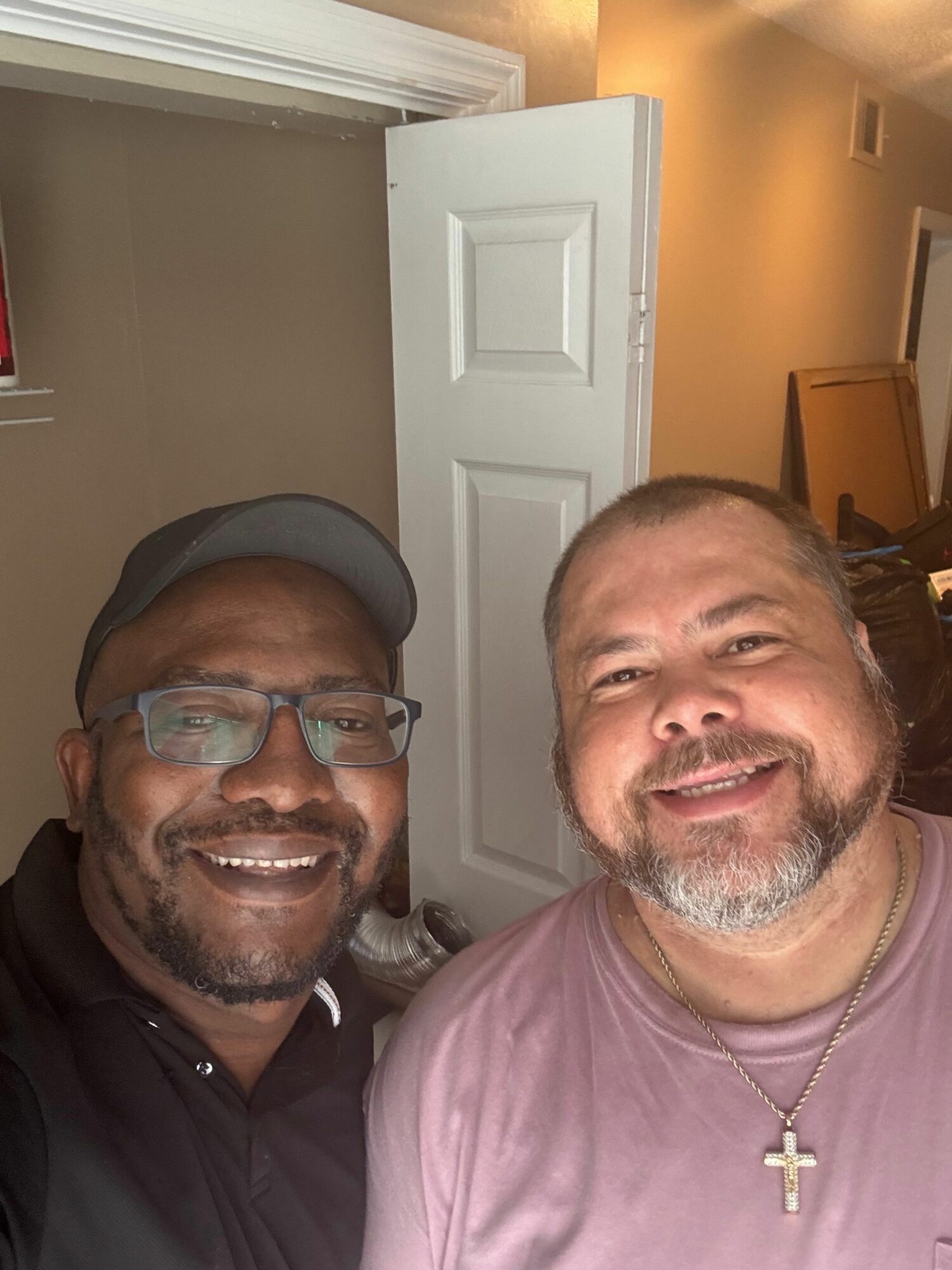 Two smiling men taking a selfie indoors, one wearing glasses and a cap, the other with a cross necklace.