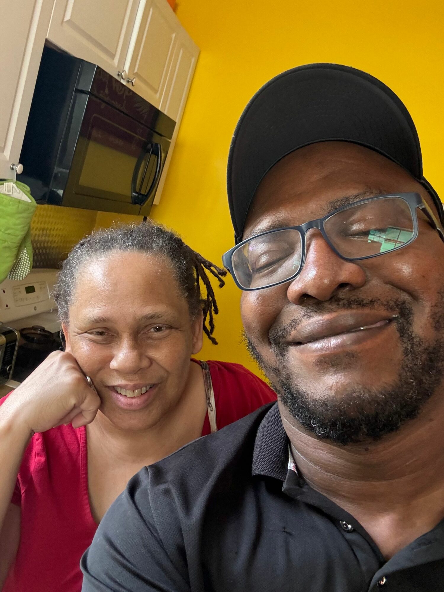Two people smiling indoors, one wearing glasses and a cap, the other with short curly hair, yellow wall background.