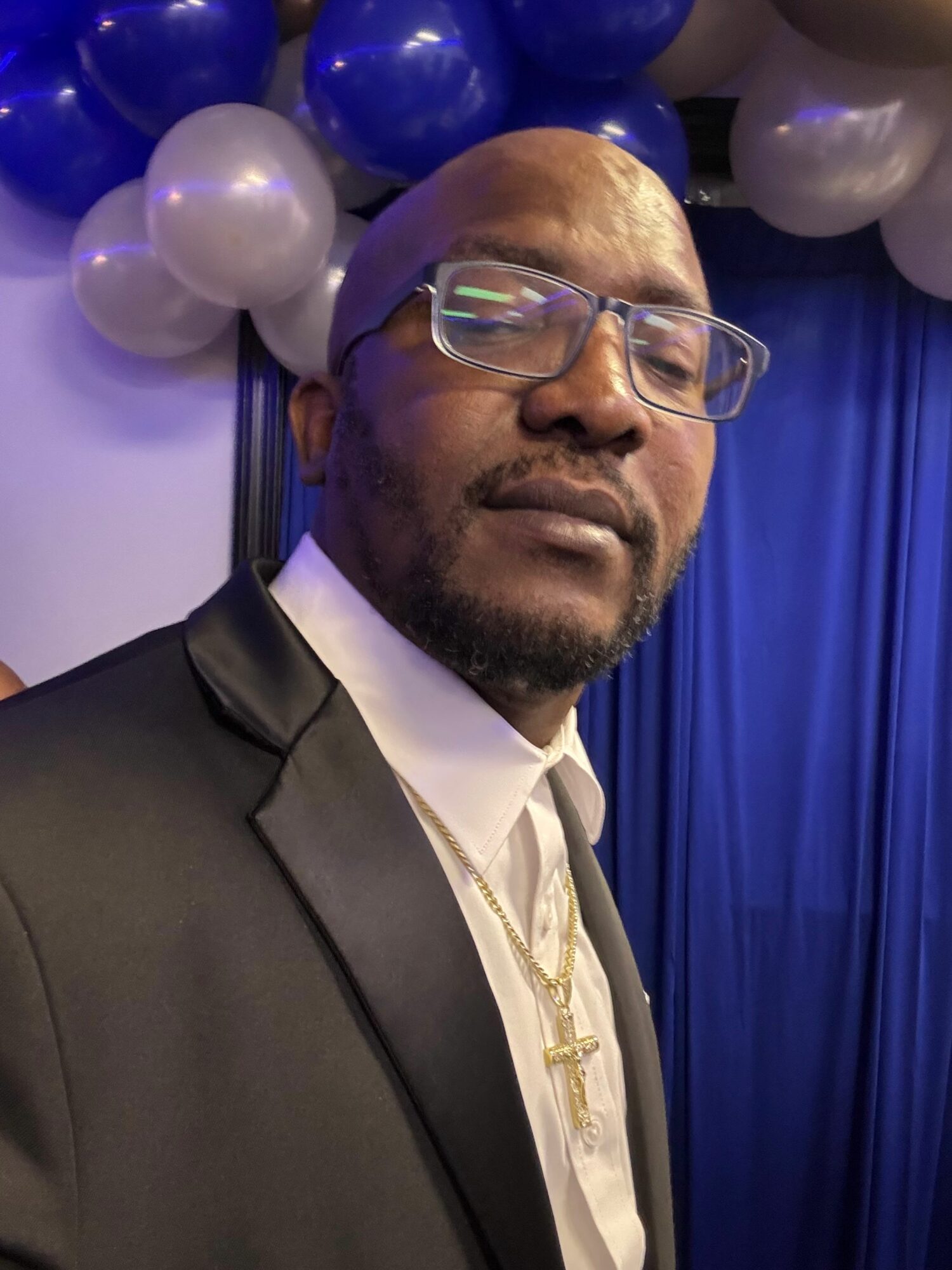 Man with glasses, beard, and gold chain, wearing a suit and tie, standing in front of blue and white balloons.