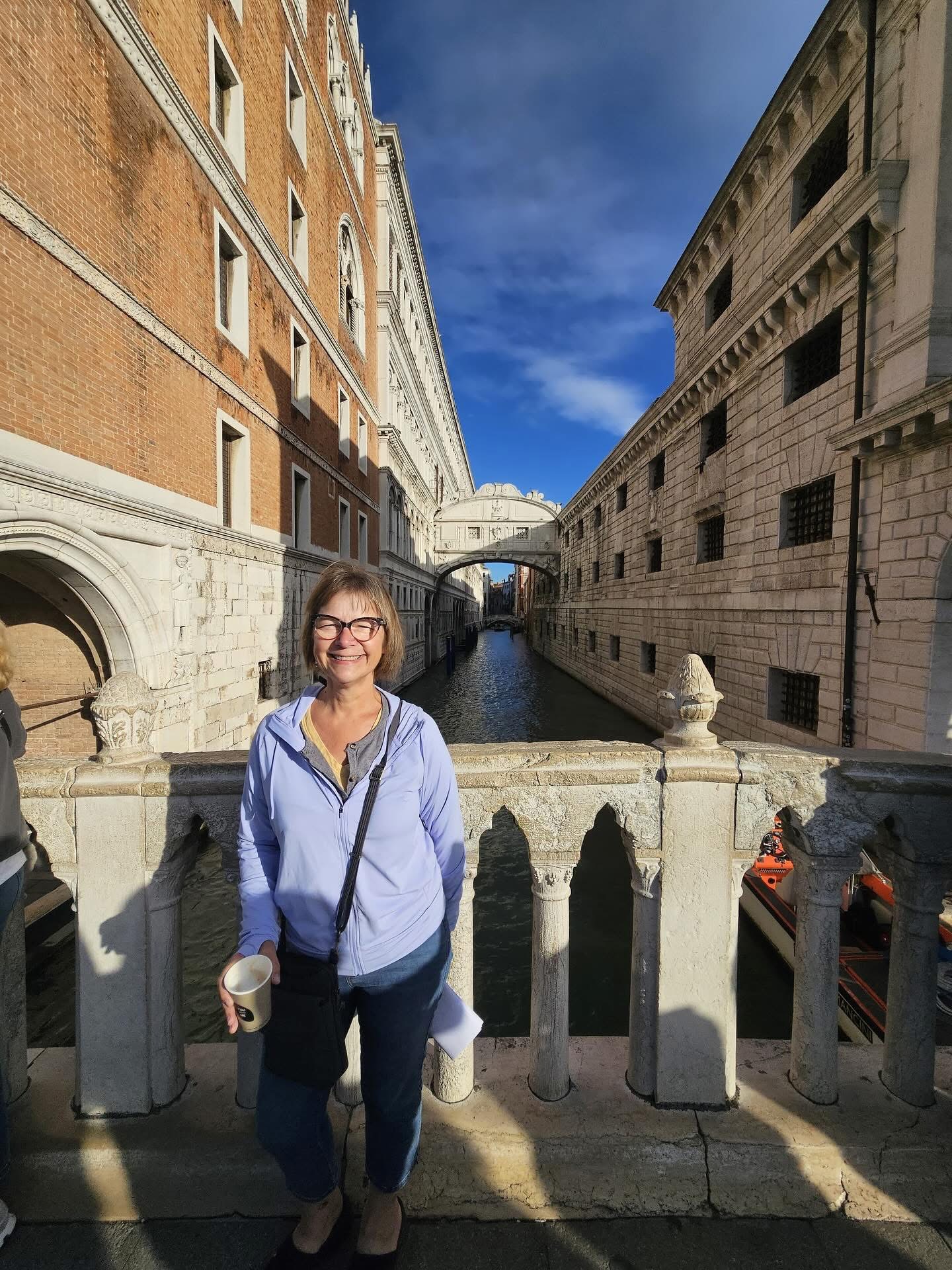 Woman with glasses and a light blue jacket standing on a bridge in Venice, Italy, with historic buildings and canal behind her.