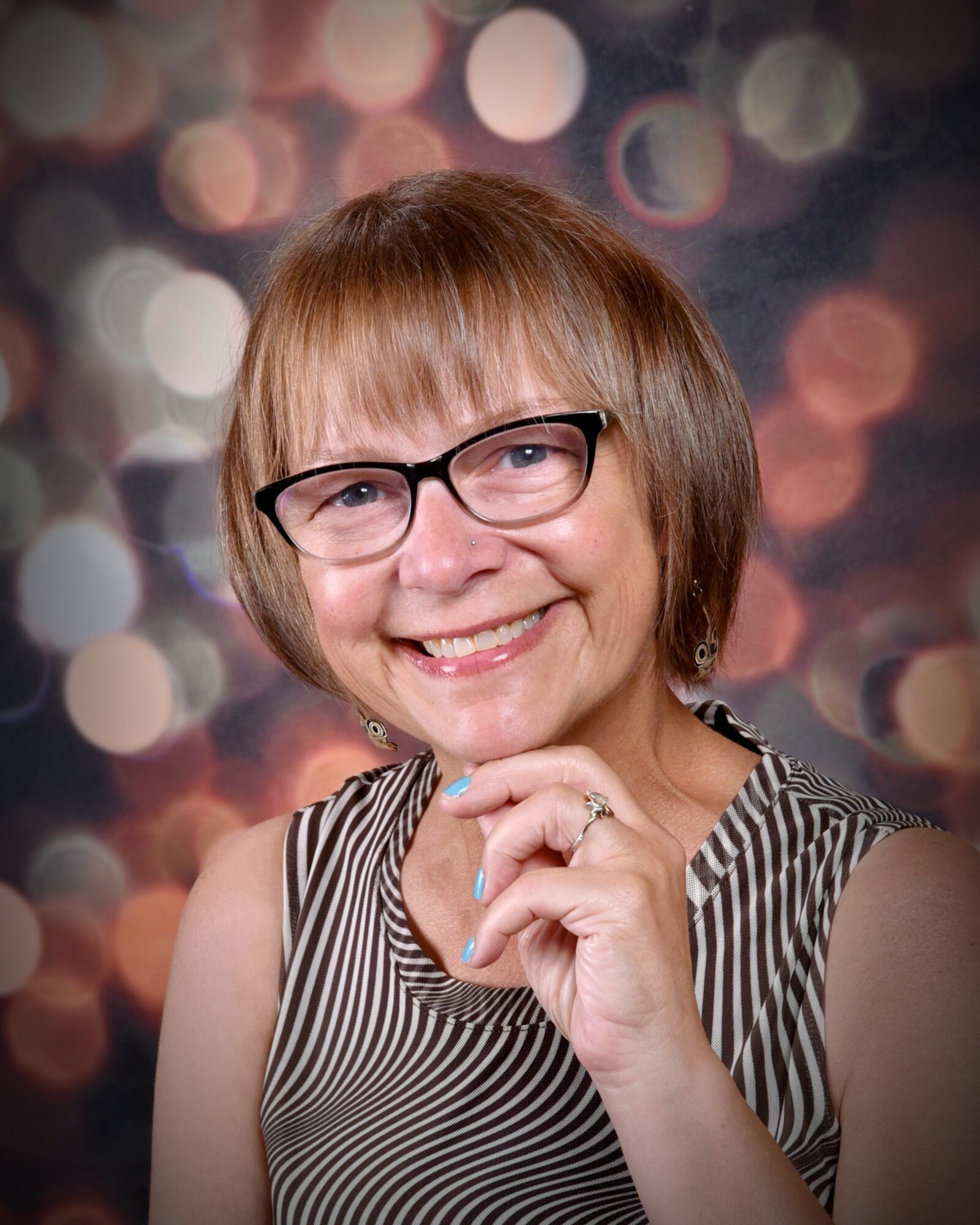 Smiling woman with glasses and short hair, wearing a striped sleeveless top, against a blurred bokeh background.