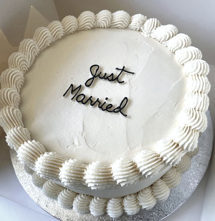 Round wedding cake with white frosting and piped border, inscribed with black cursive 'Just Married'.