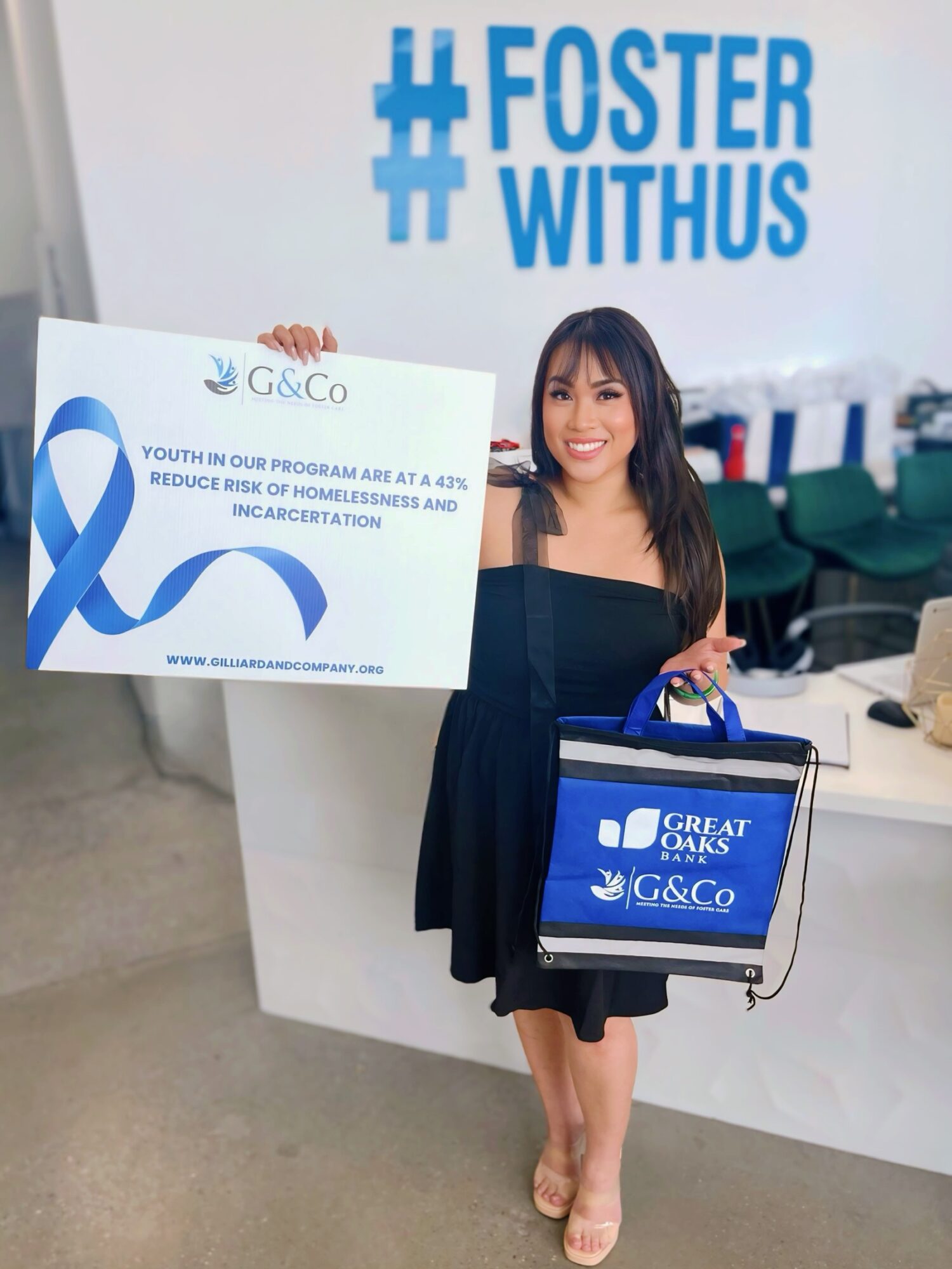 Young woman with dark hair holding a box and standing in front of a white wall with blue text and logo.