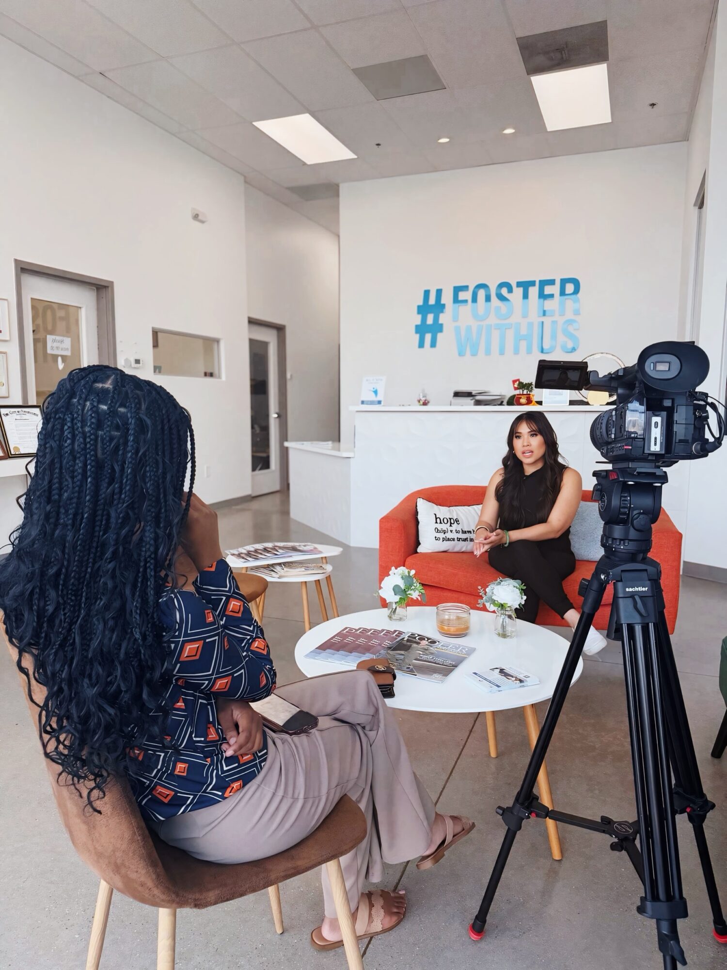 Woman sitting at a table in a room with a camera recording her, hashtag on wall behind her, and various items on table.