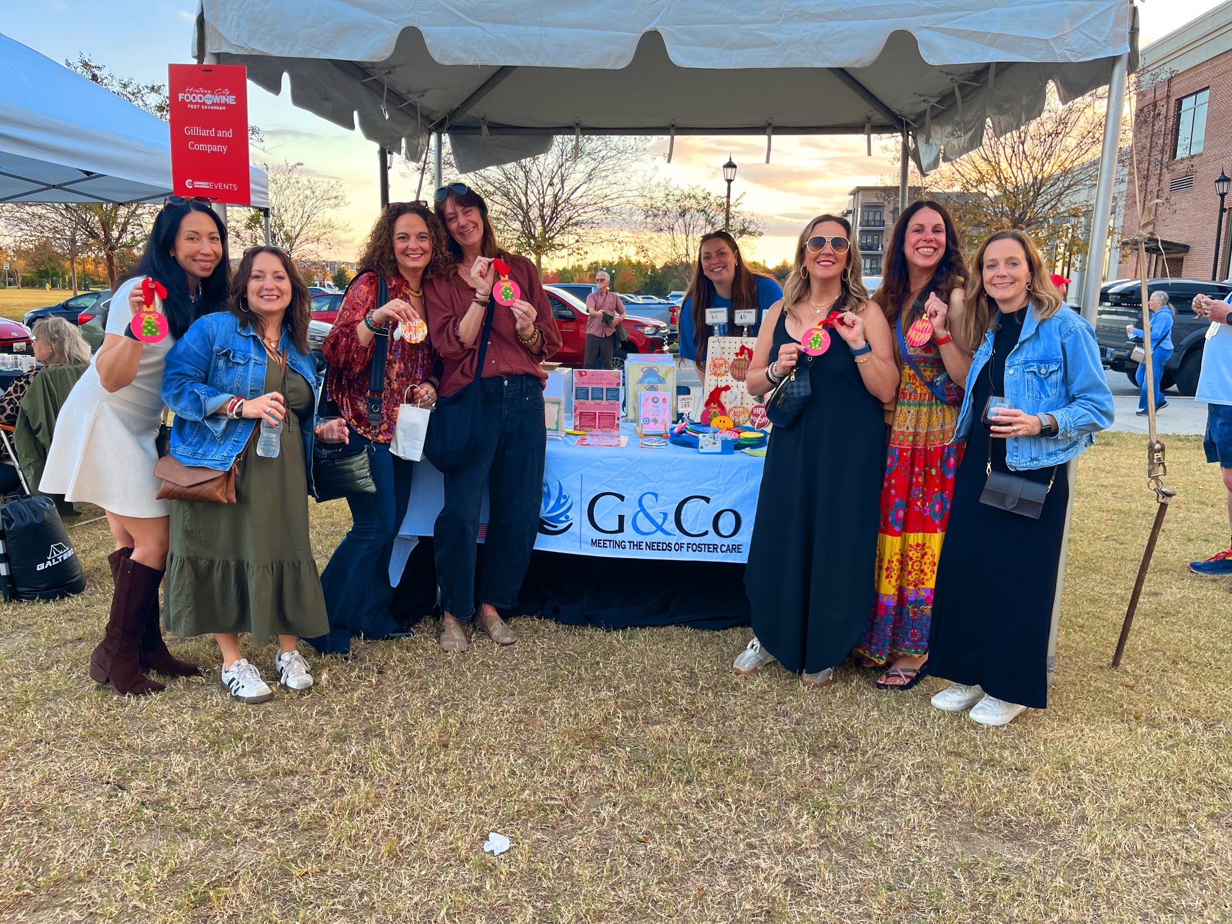 Group of eight women standing outdoors under a tent, smiling, with a table and buildings in the background.