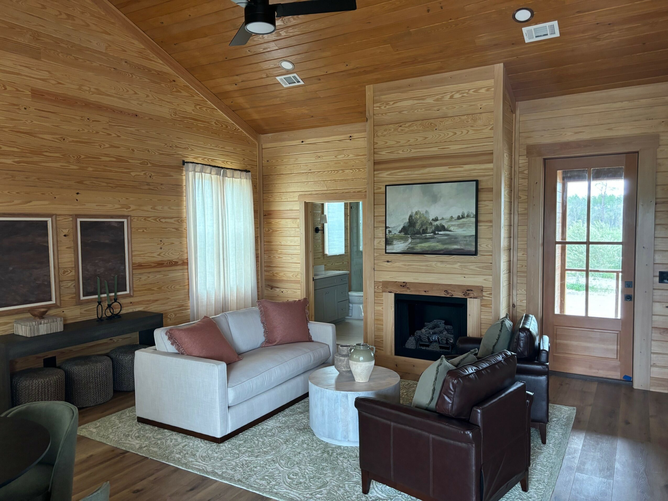 Living room with wooden walls and ceiling, white sofa with pink pillows, dark armchair, fireplace, window, and door.