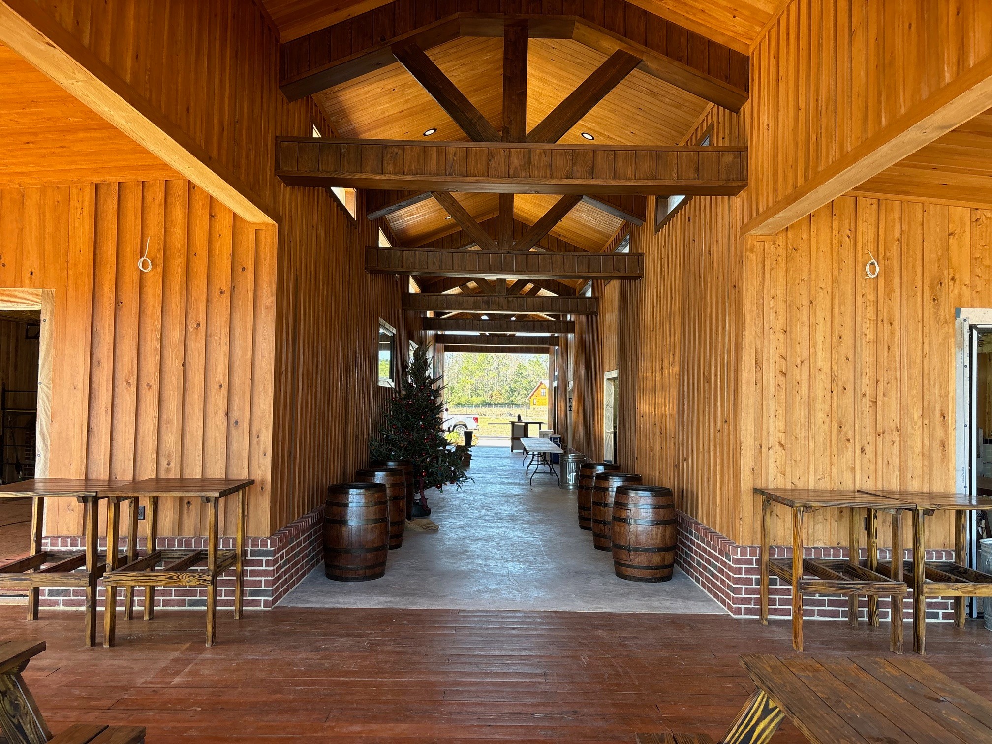 Wooden corridor with tables and barrels, leading to a bright outdoor area with trees and a bench.