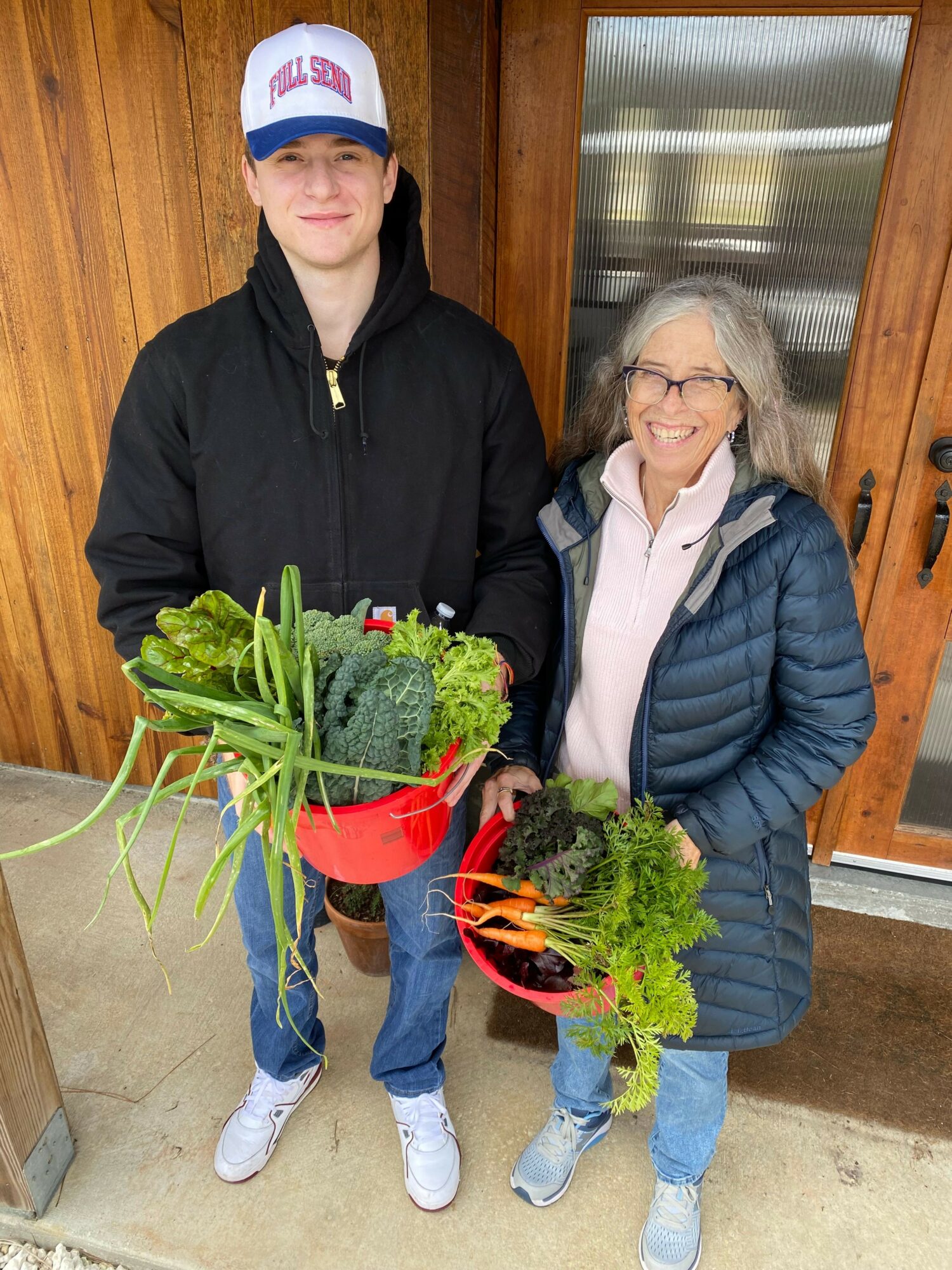 Young person and older woman holding baskets of fresh vegetables indoors, smiling, with wooden walls and glass door behind.