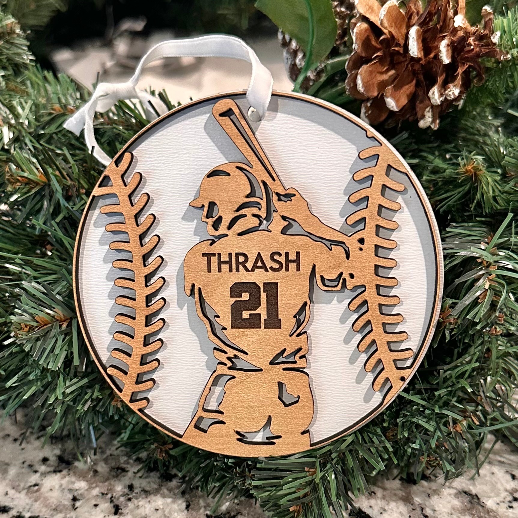Decorative ornament with a baseball player silhouette, surrounded by pine branches and pinecones, with a white ribbon at the top.