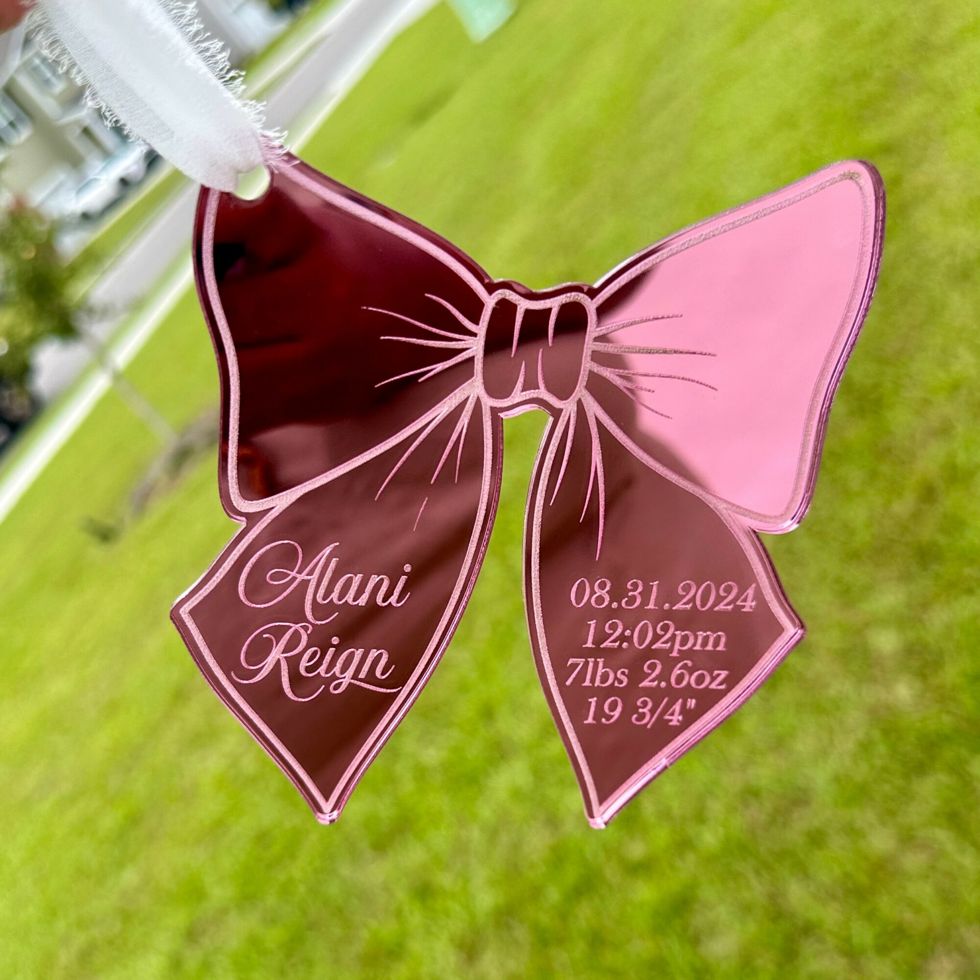 Pink and burgundy bow-shaped ornament with birth details and name, on a grassy background.