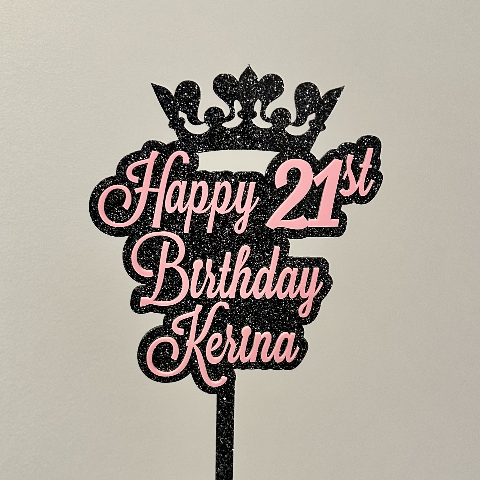 Black glittery birthday sign with pink text reading 'Happy 21st Birthday Kerina' and a crown on top.