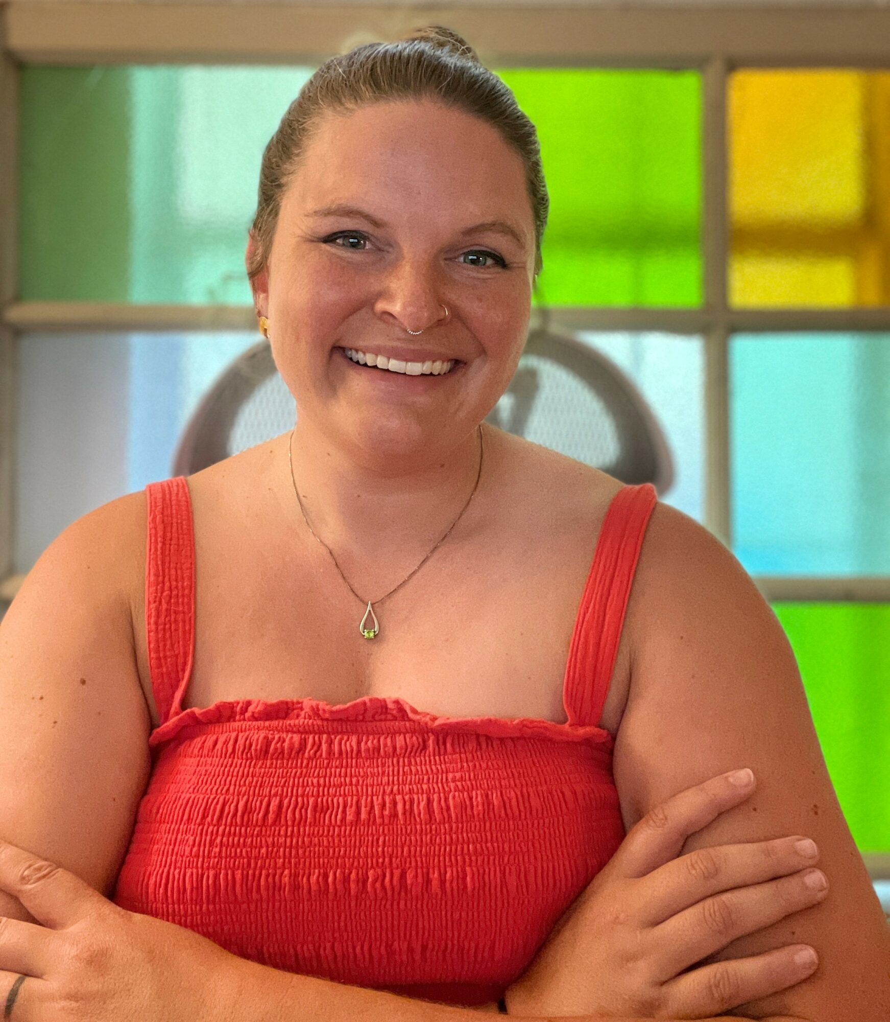 Smiling woman with light brown hair in a bun, wearing a red sleeveless top and a necklace, standing in front of colorful stained glass windows.