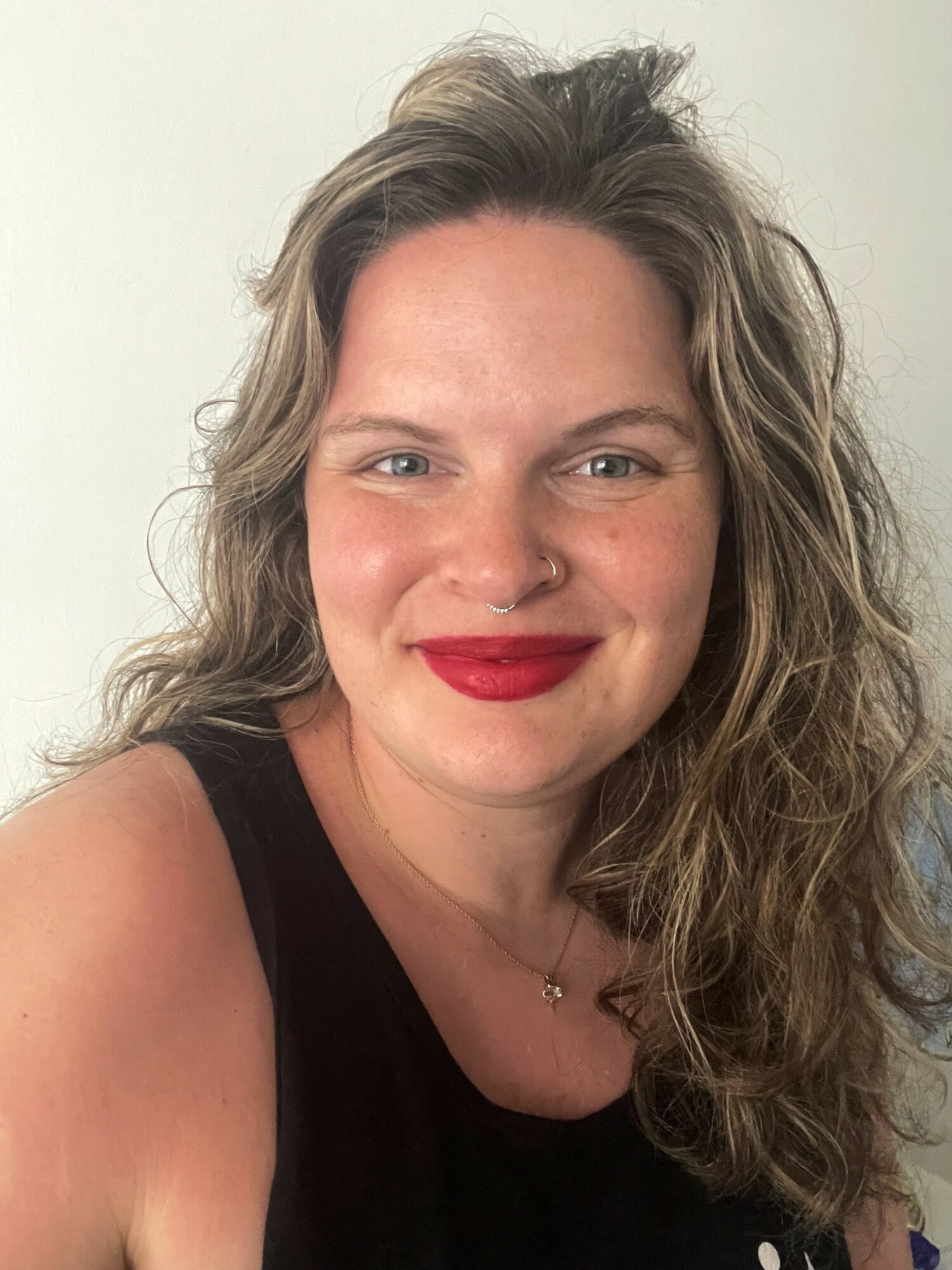 Woman with wavy hair, red lipstick, and a nose ring smiling at the camera, wearing a black top.