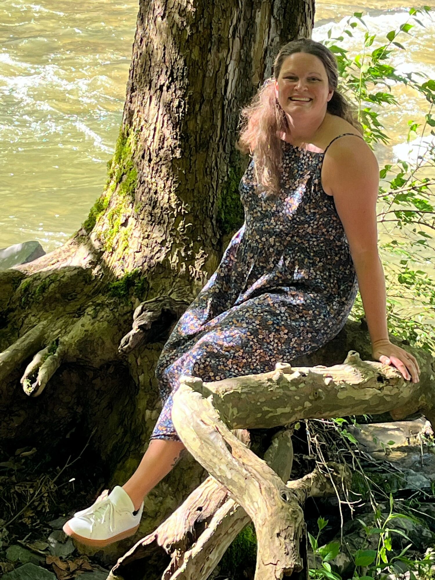 Woman sitting on a tree branch outdoors, surrounded by green foliage, wearing a patterned dress and white shoes.